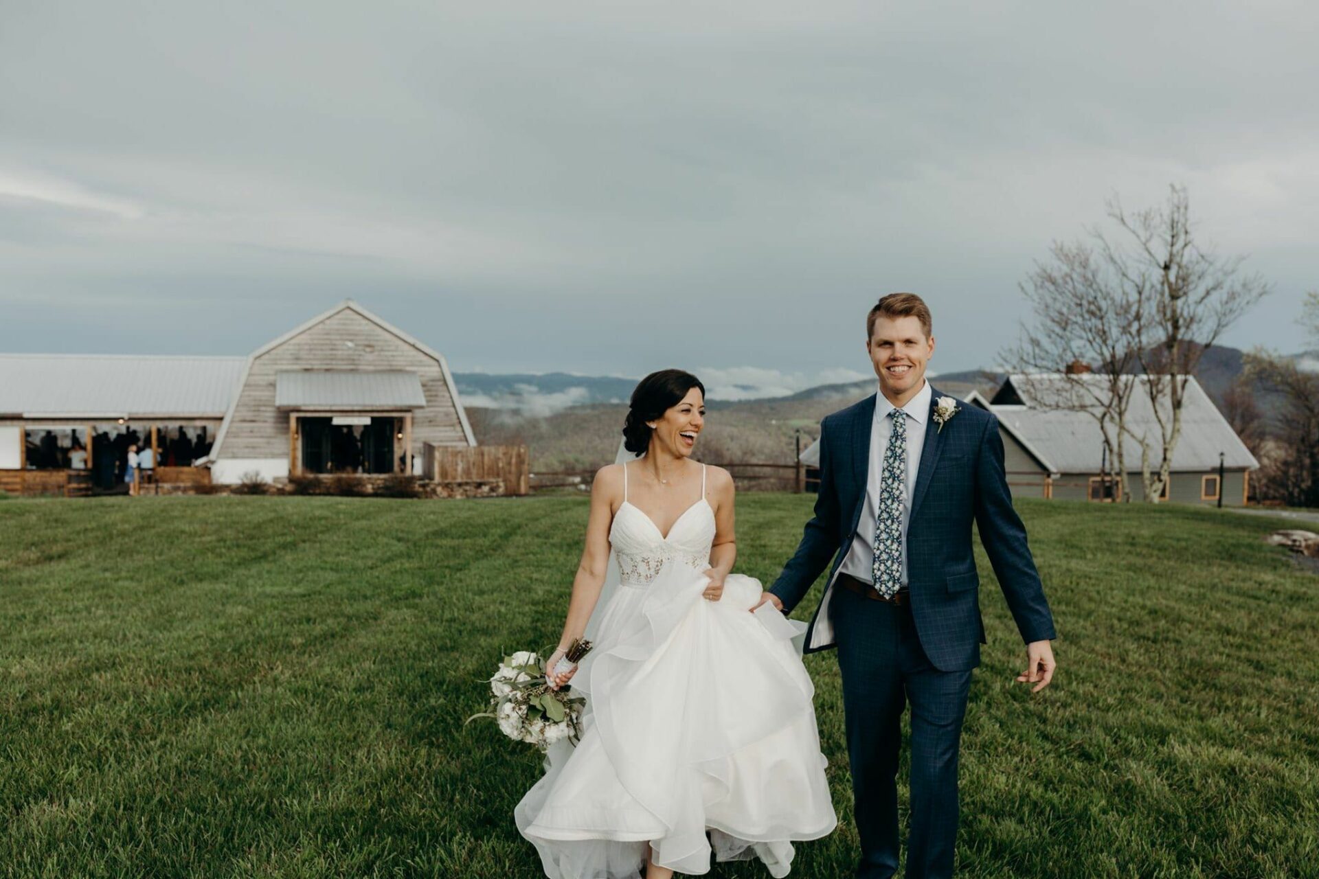 A bride and groom walking through a grassy field with a barn in the background.