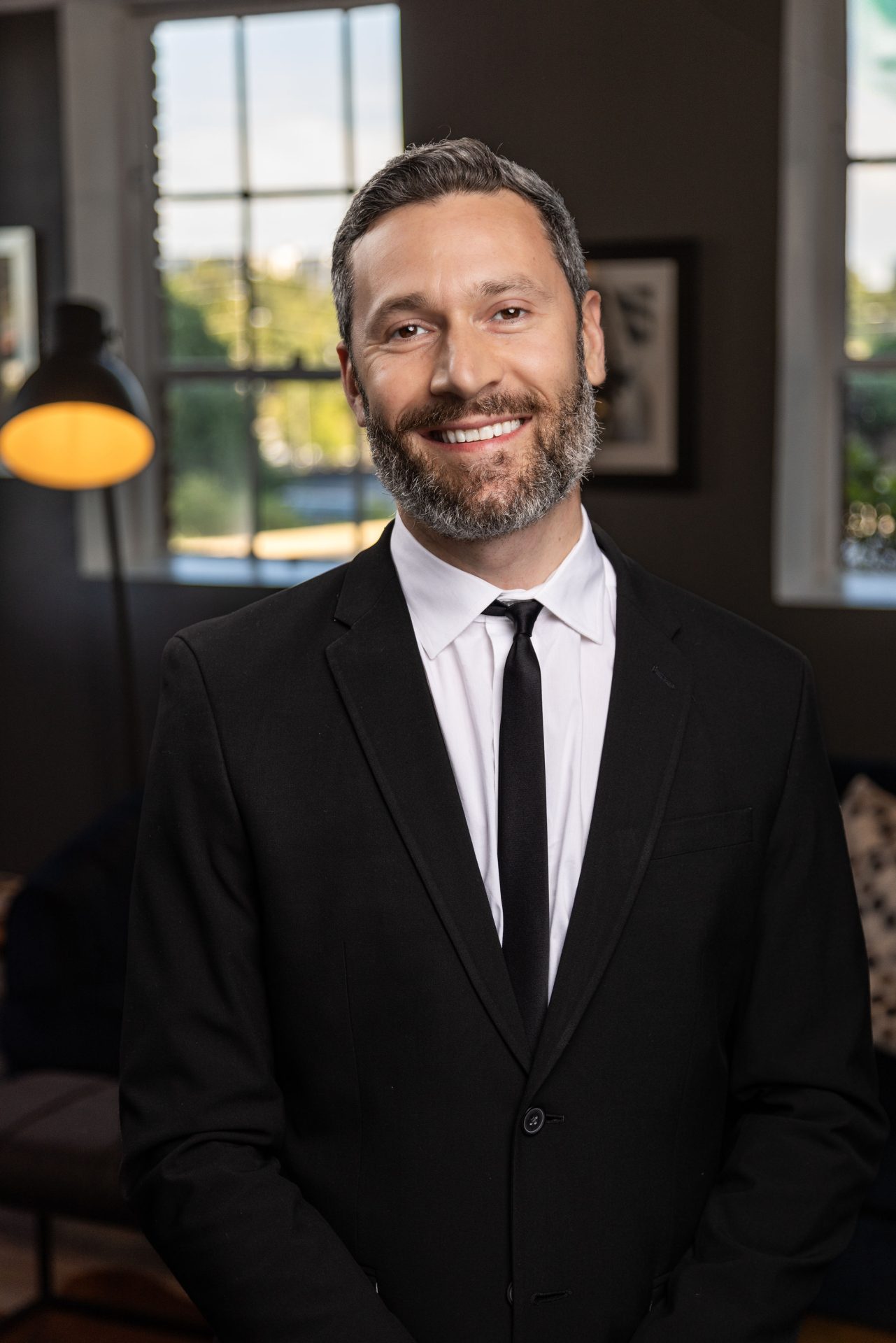 A man in a black suit and tie smiles while standing indoors with a blurred window and lamp in the background.
