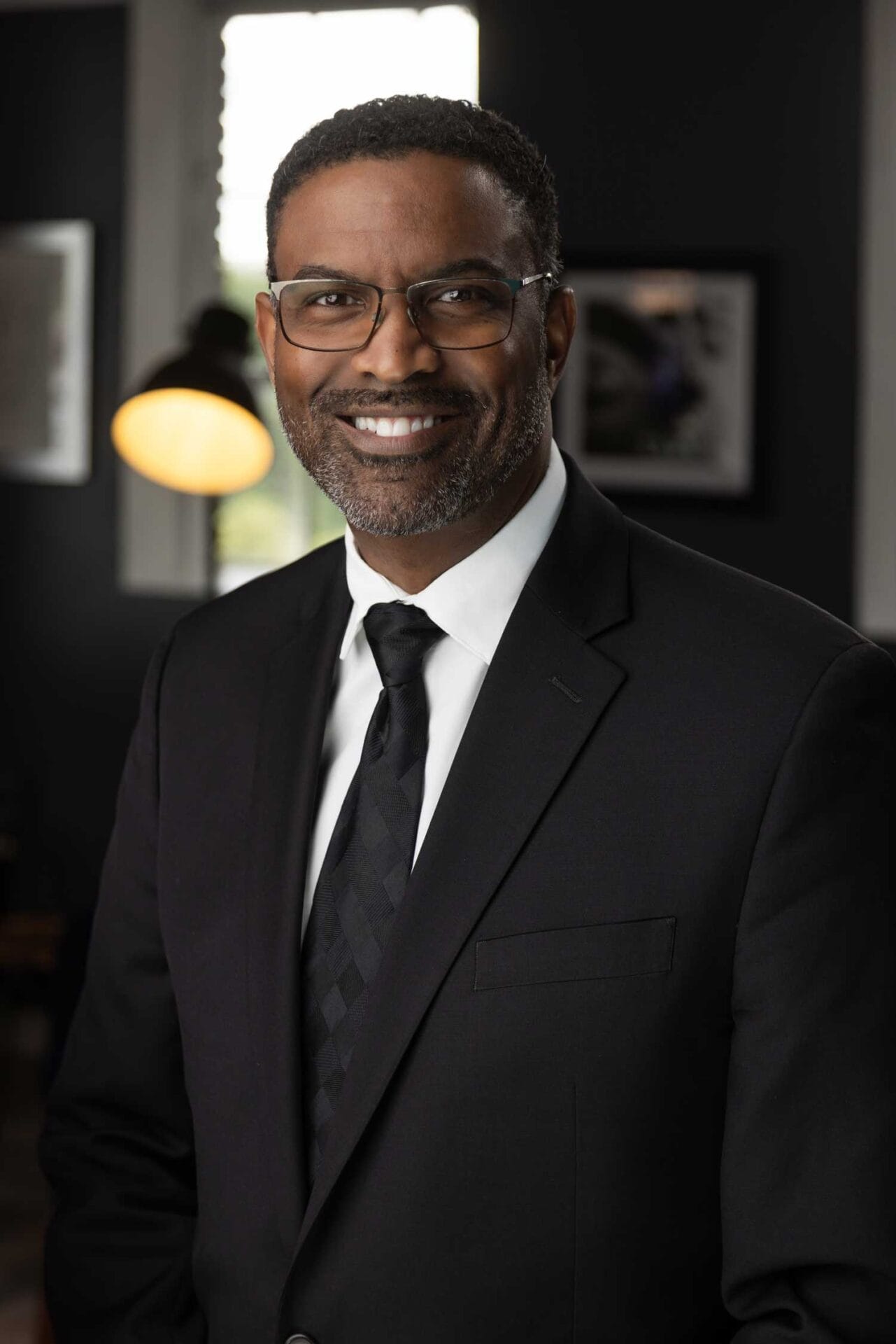 A man in a dark suit, white shirt, and dark tie stands indoors, smiling at the camera. He has short hair, a trimmed beard, and wears glasses.