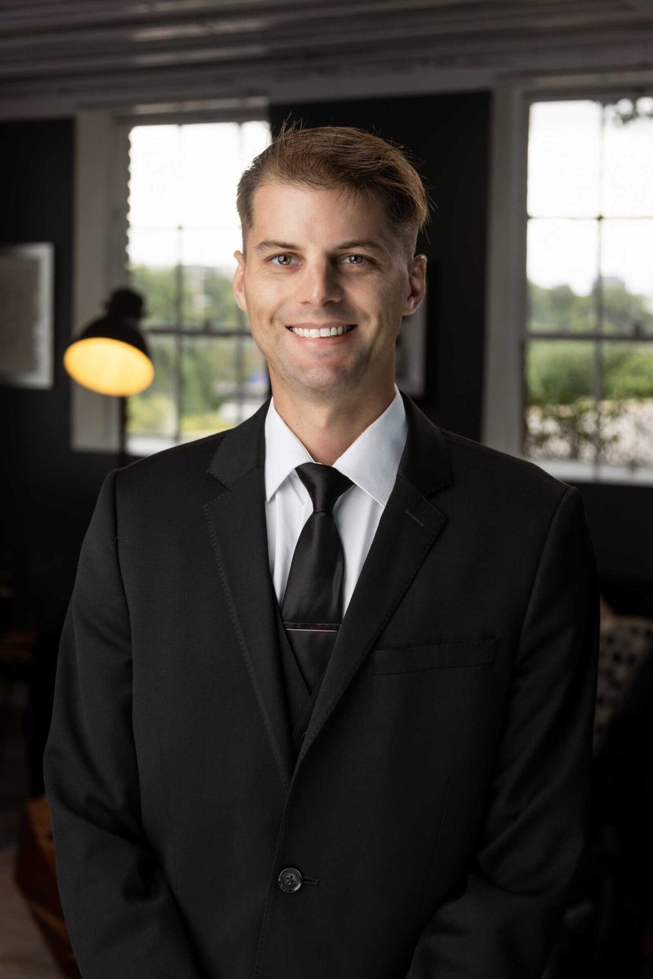 A man in a black suit, white shirt, and black tie stands indoors, smiling at the camera with windows and a lamp in the background.