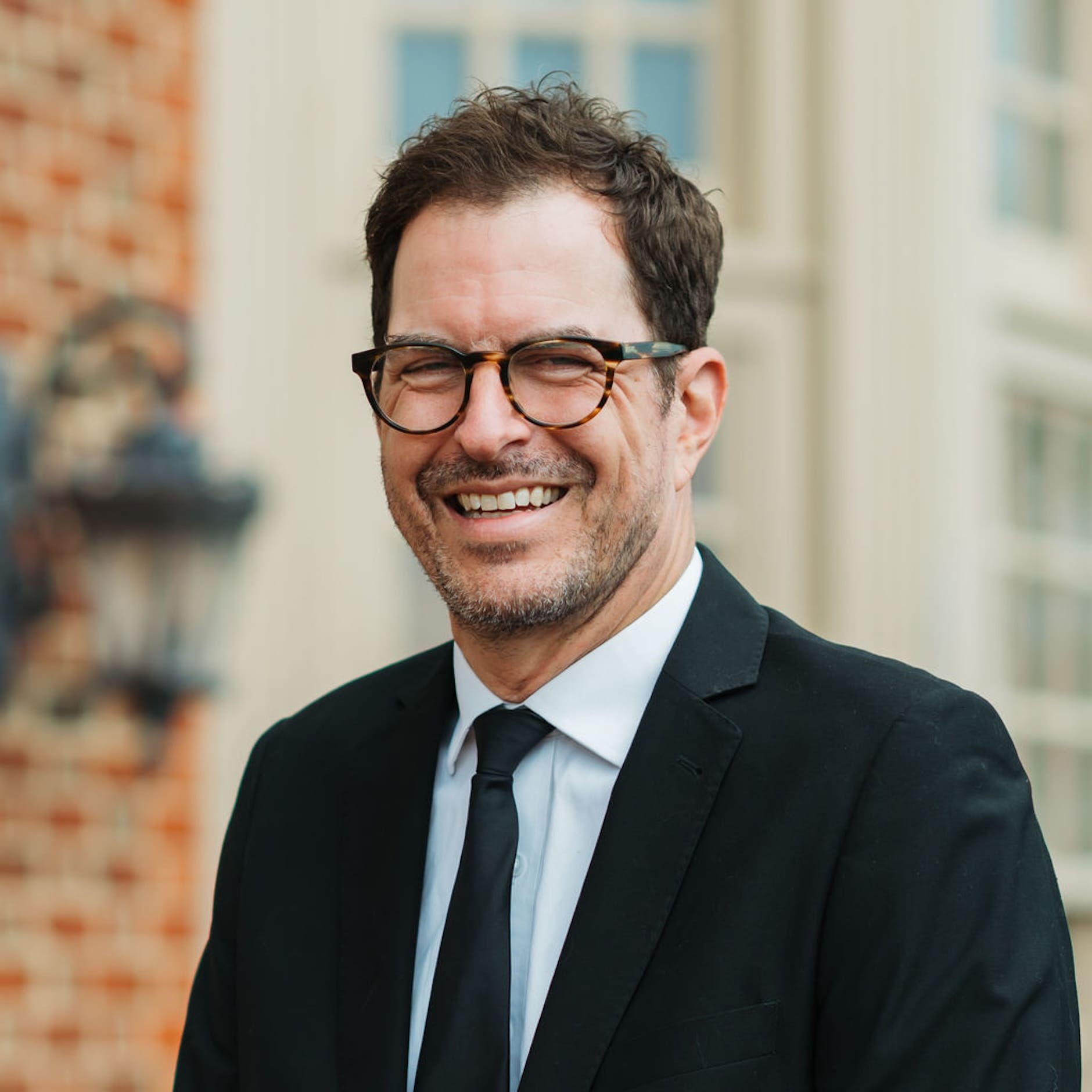 Cam D Headshot 2 A man in a suit and tie wearing glasses is smiling outdoors in front of a building with brick and white details.
