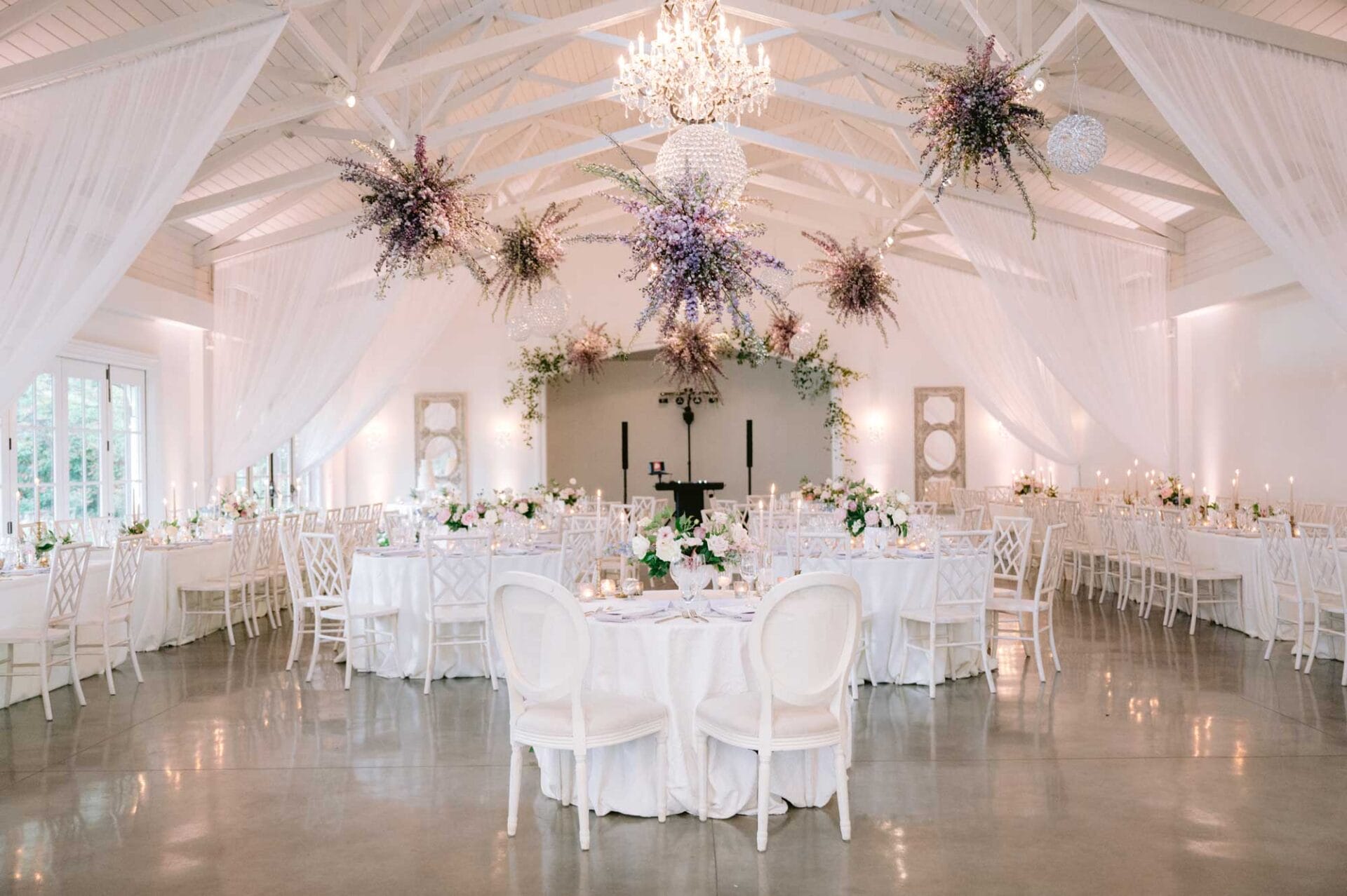 Elegant banquet hall with round tables covered in white linens, floral centerpieces, white chairs, hanging floral arrangements, and a chandelier under a vaulted white ceiling.