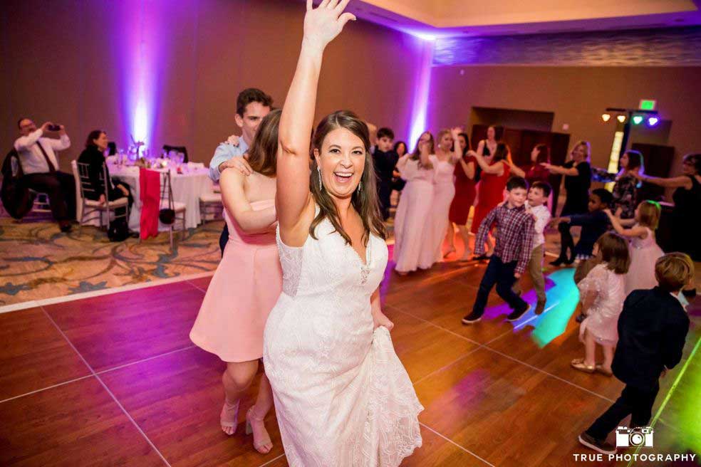 Cape-Rey-Wedding A woman in a white dress smiles and raises her hand while dancing at a lively indoor event with adults and children on the dance floor.