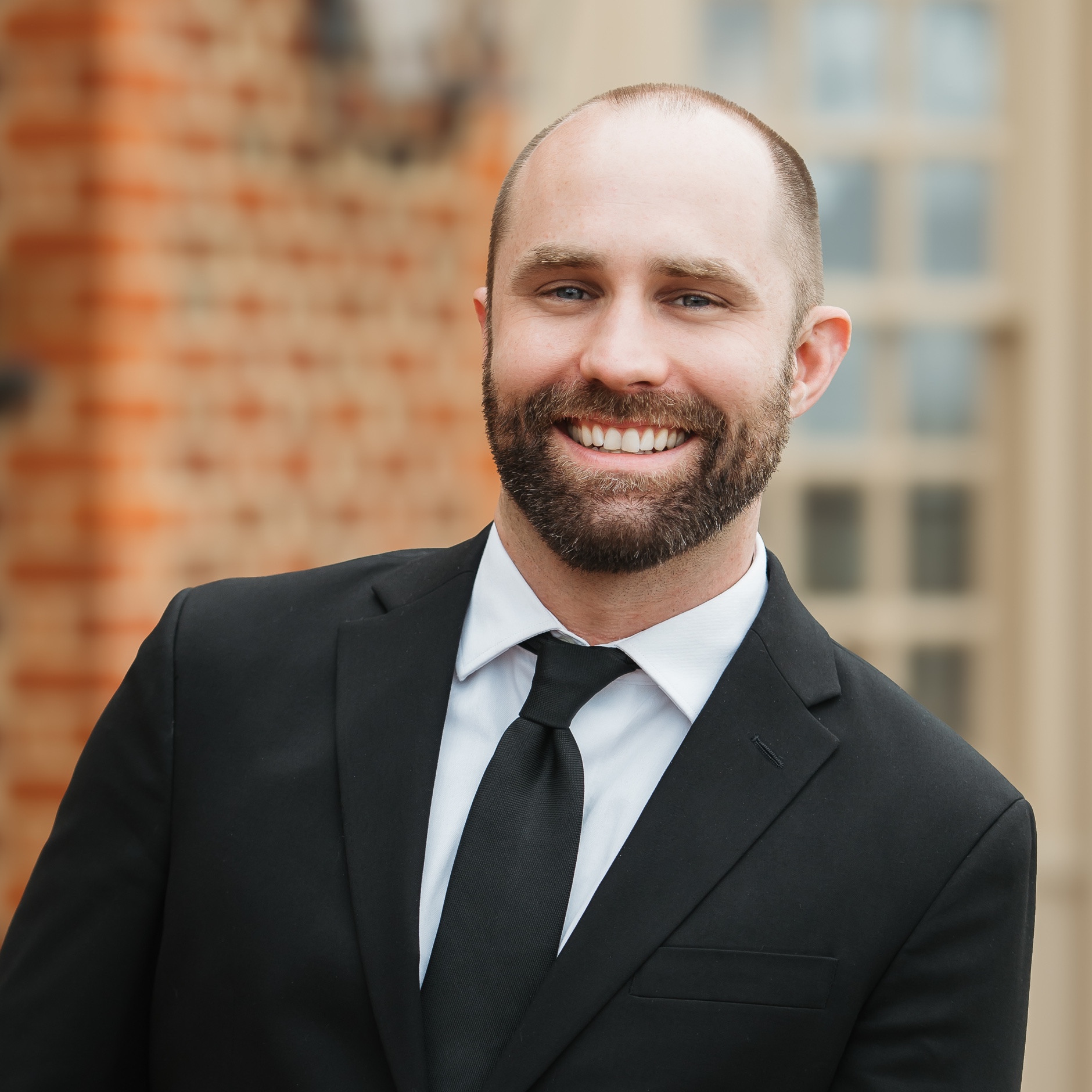 Chris 3 copy A man in a suit and tie smiling in front of a brick building.