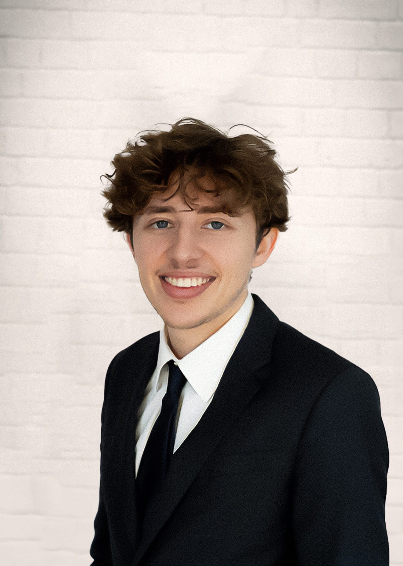 A young man with curly brown hair in a black suit and tie smiling in front of a white brick wall.