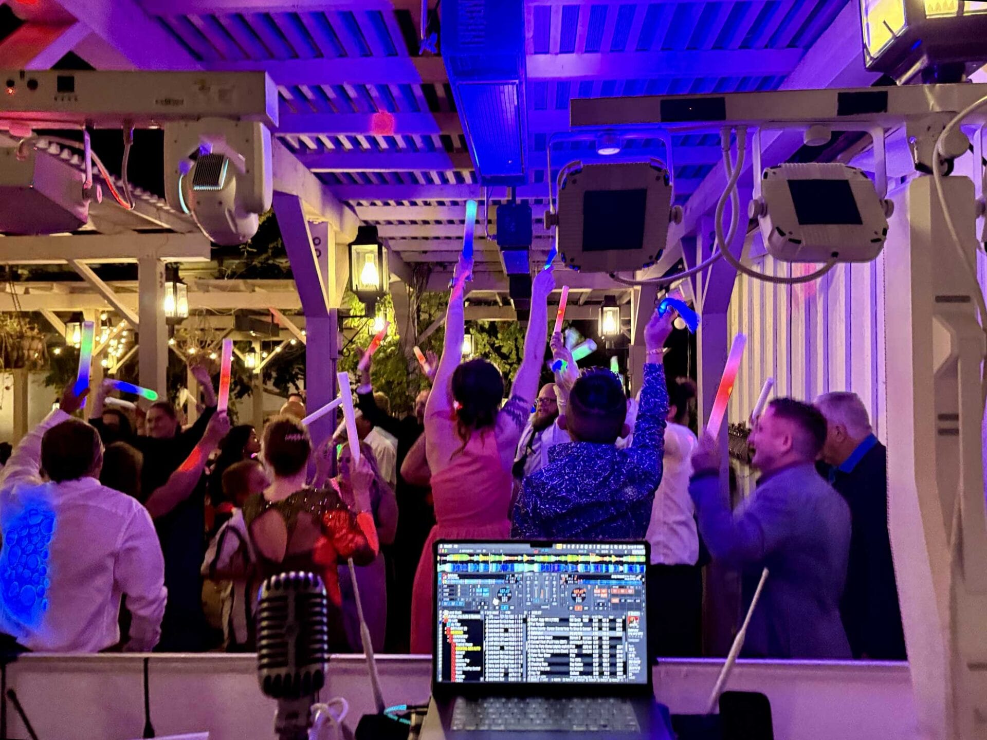 Cosmopolitan-Dance-Floor A group of people at a lively indoor party holding glow sticks, seen from behind a DJ booth with a laptop and microphone in the foreground.