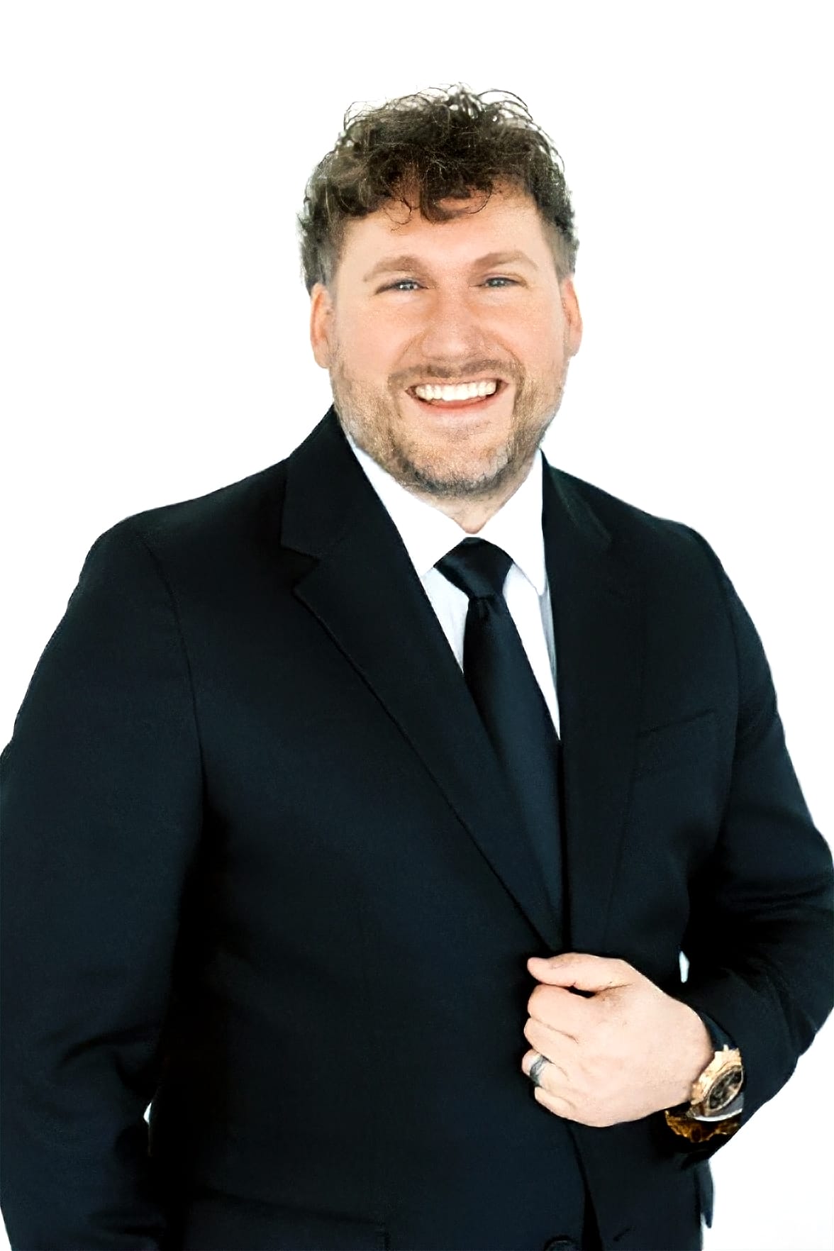 DJ-Jeff_Stevens Man in a black suit, white shirt, and black tie smiles at the camera against a plain white background. He has light skin, curly brown hair, and a beard, with one hand adjusting his jacket.
