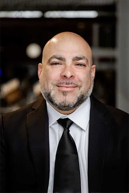 A man in a black suit, white shirt, and black tie is smiling at the camera against a blurred indoor background.