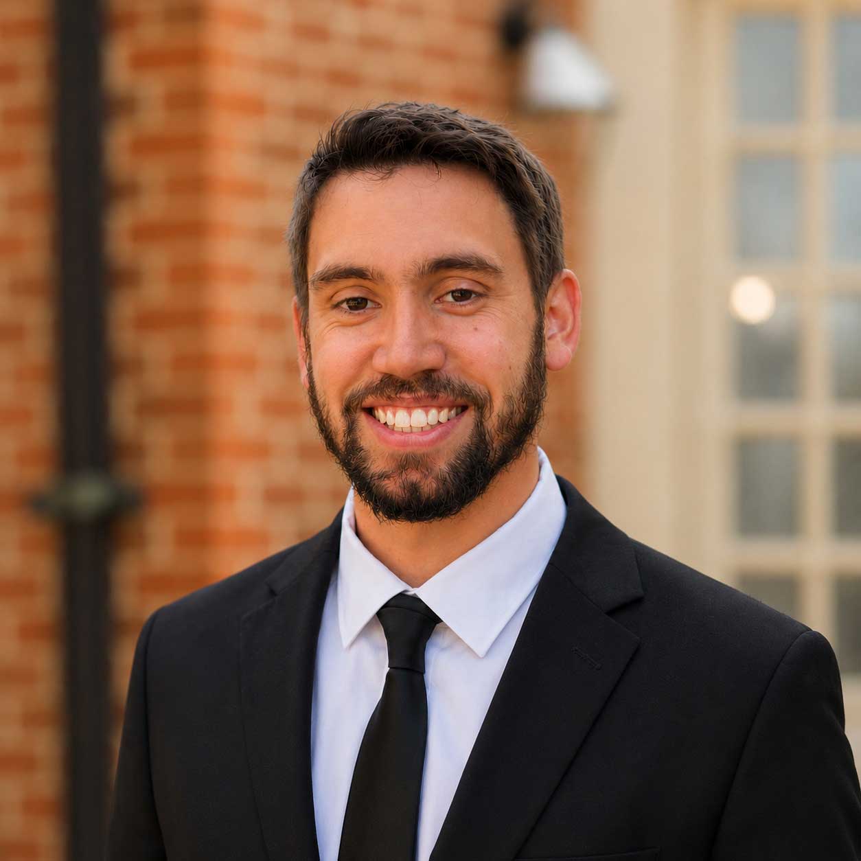 Man with short brown hair and beard wearing a black suit, white shirt, and black tie, standing outdoors in front of a building with brick and glass.