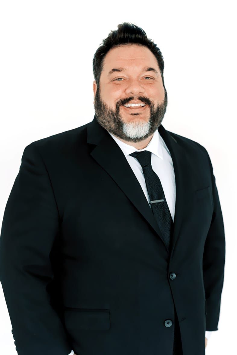DJ-brandon_Alley Man with dark hair and beard wearing a black suit, white shirt, and black tie, standing and smiling against a plain white background.