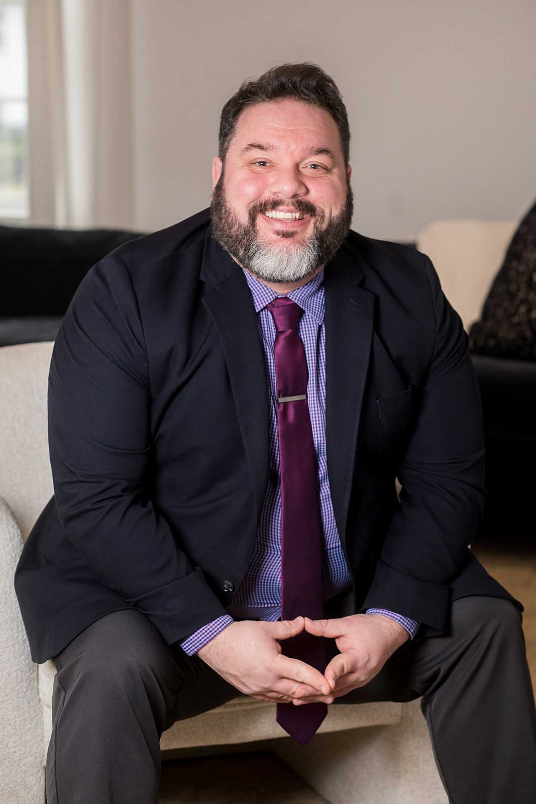 DJ_Bunn-Charlotte-Brandon A man with a beard and mustache, wearing a dark suit and purple tie, sits on a light-colored chair and smiles at the camera.