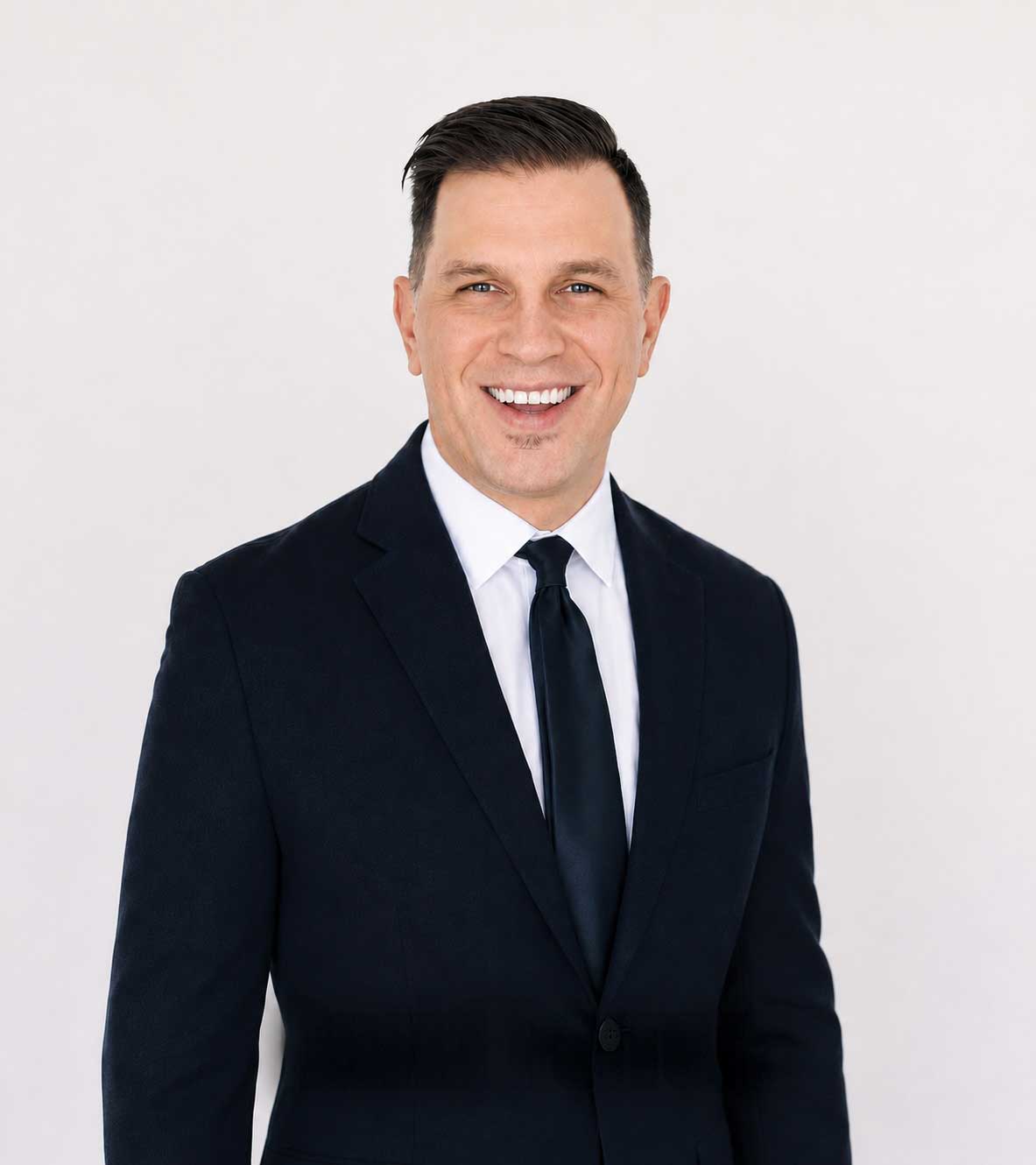 Man in a dark suit, white shirt, and dark tie standing against a plain light background, smiling at the camera.