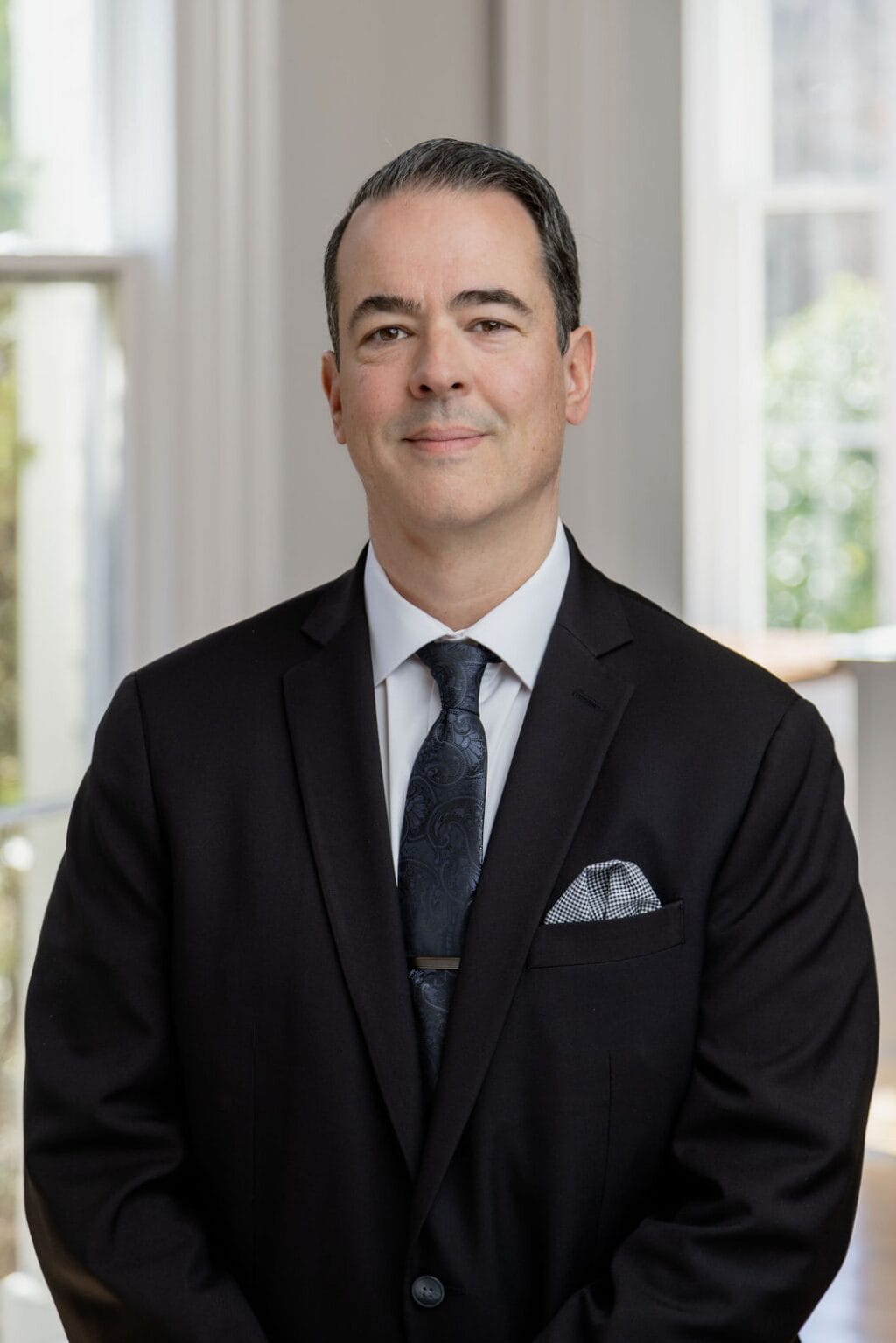 A man in a black suit and tie stands indoors, smiling slightly, with a window in the background.