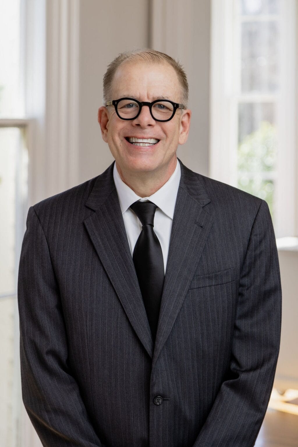 Man in a pinstripe suit and tie stands smiling in a bright room with windows in the background.