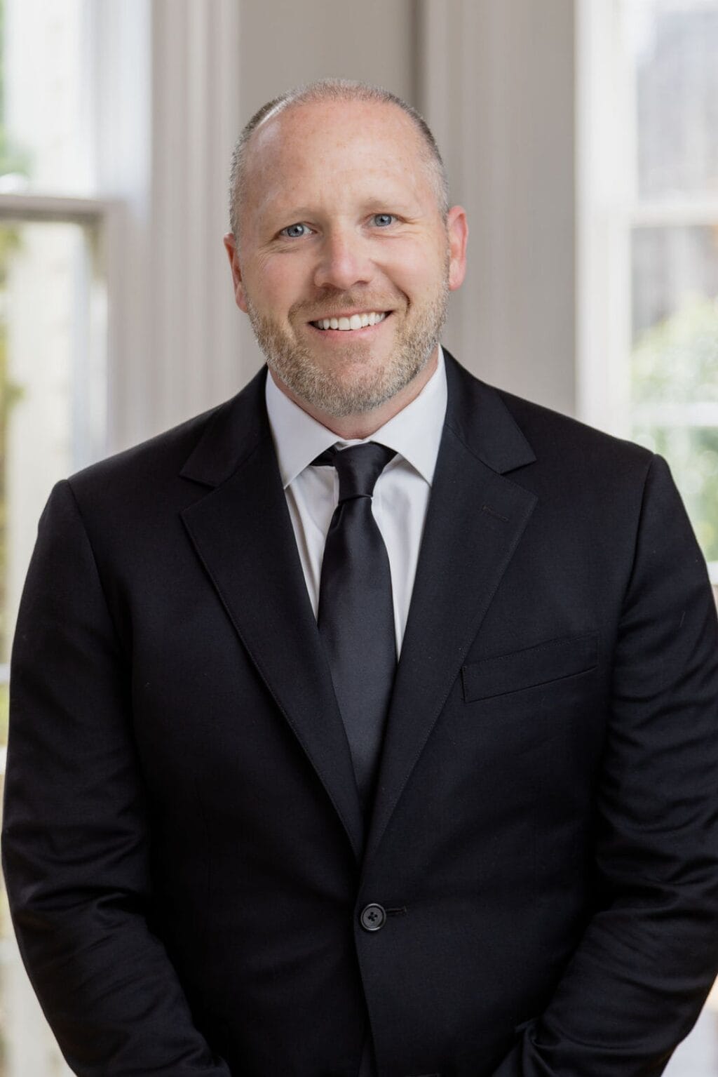 A man in a black suit and tie smiles, standing in a room with large windows.