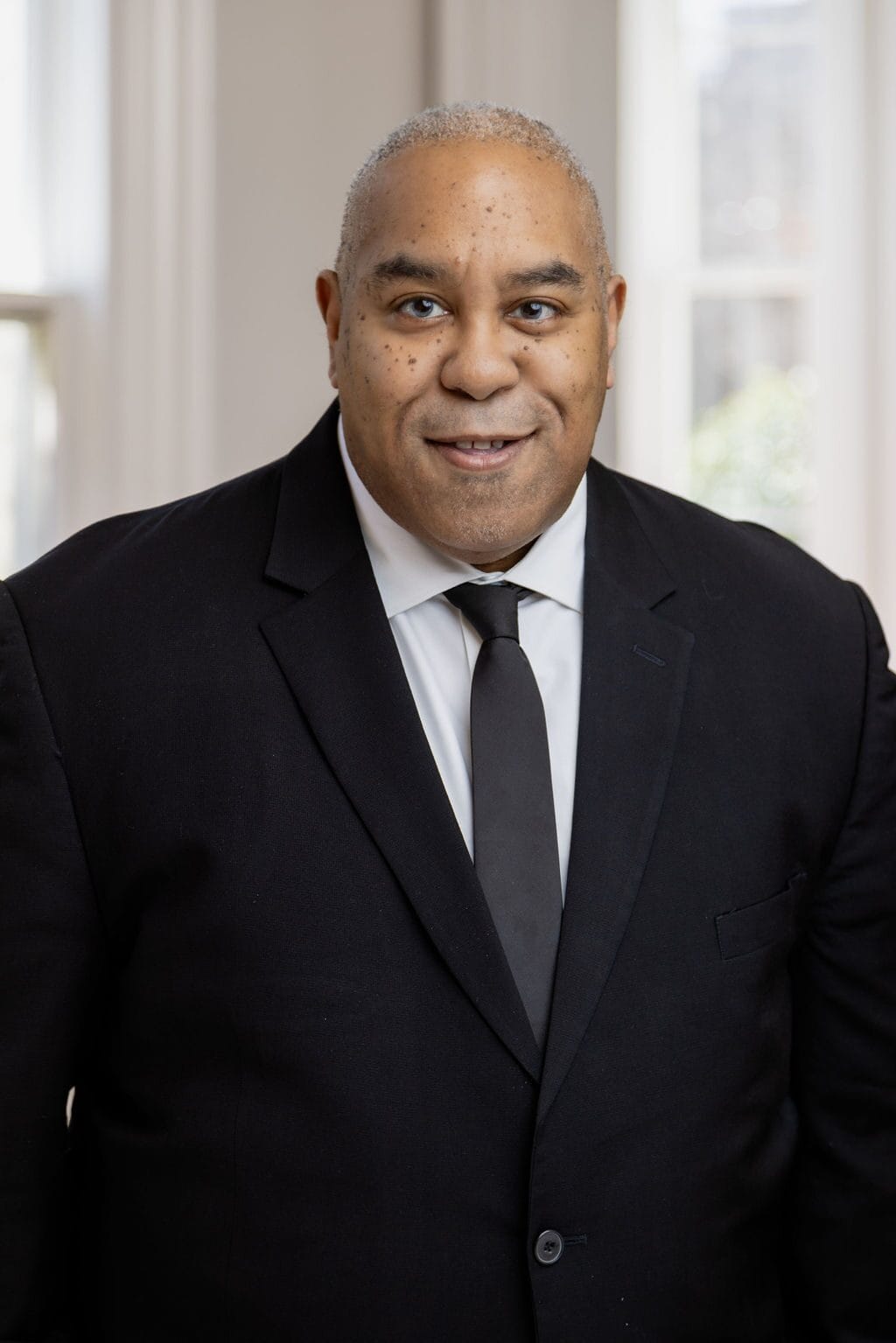 A man in a black suit and tie stands indoors, facing the camera, with a neutral expression. He has a shaved head and is positioned in front of bright windows.