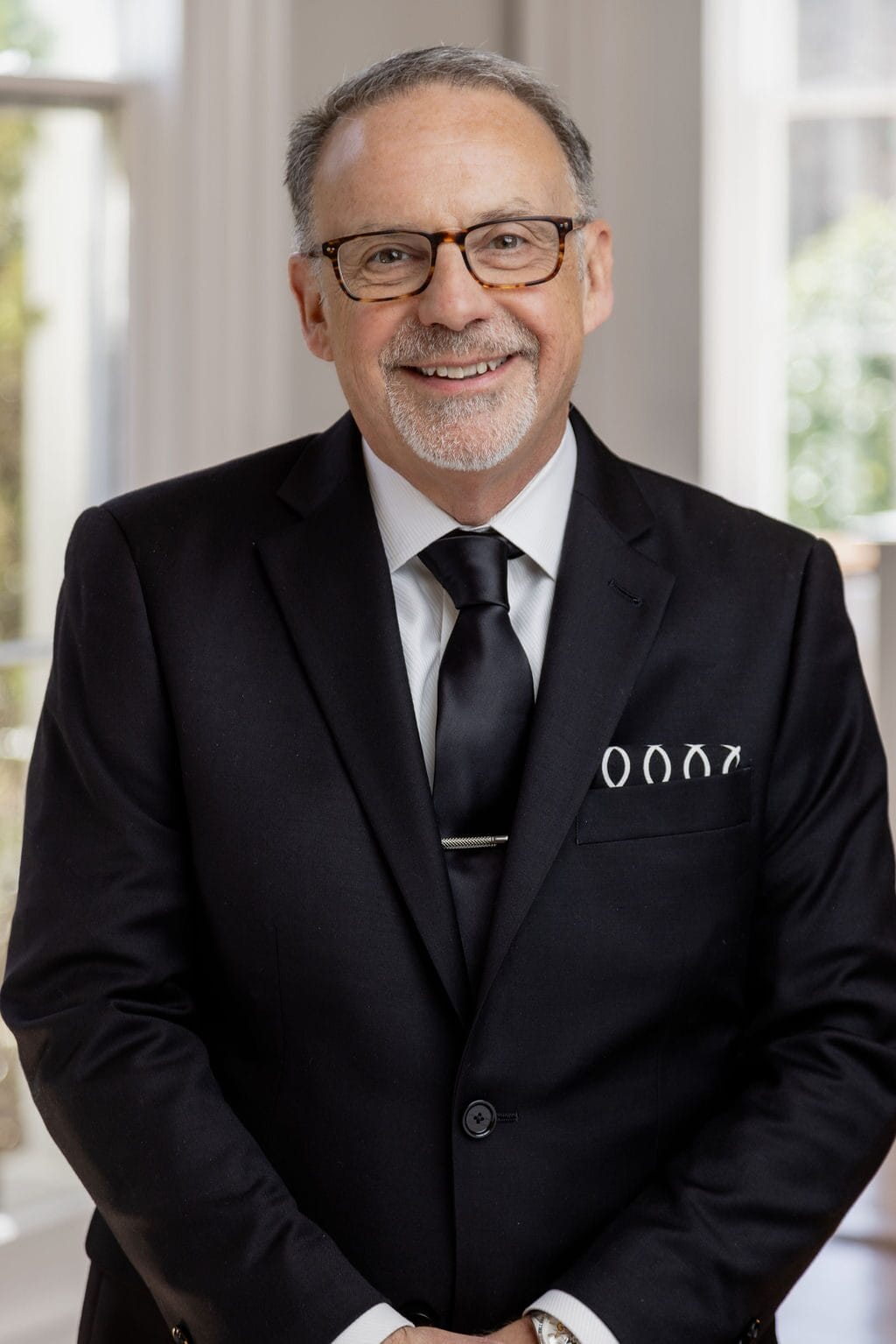 Man in a dark suit and tie, with glasses and graying hair, smiling in a well-lit room.