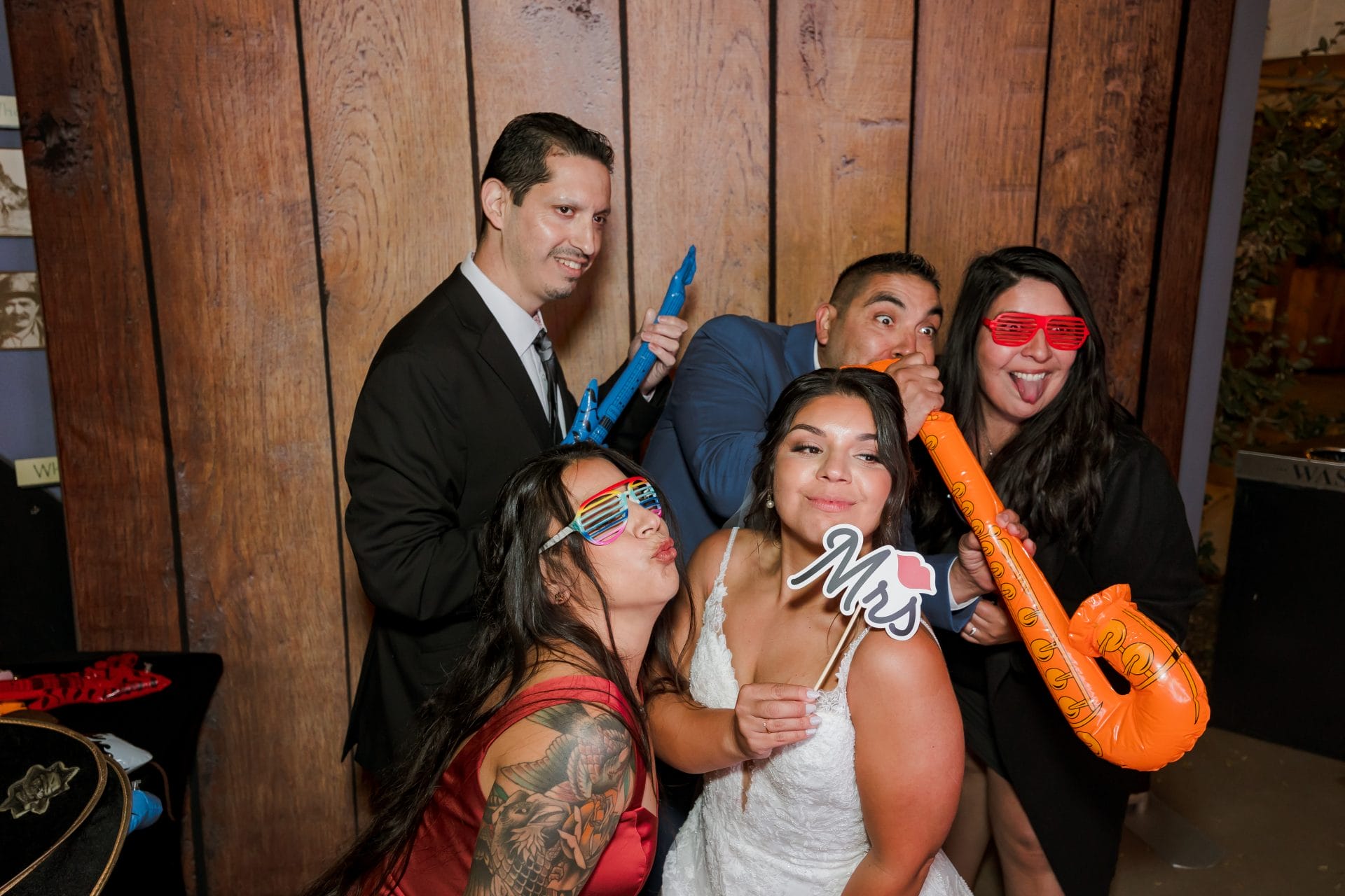 Five adults pose playfully at a photo booth, wearing novelty glasses and holding props, including inflatable instruments and a "Mrs." sign, in front of a wooden backdrop.