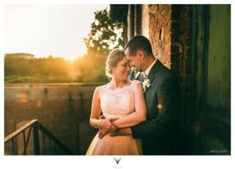 A bride and groom embrace in front of a brick building at sunset.