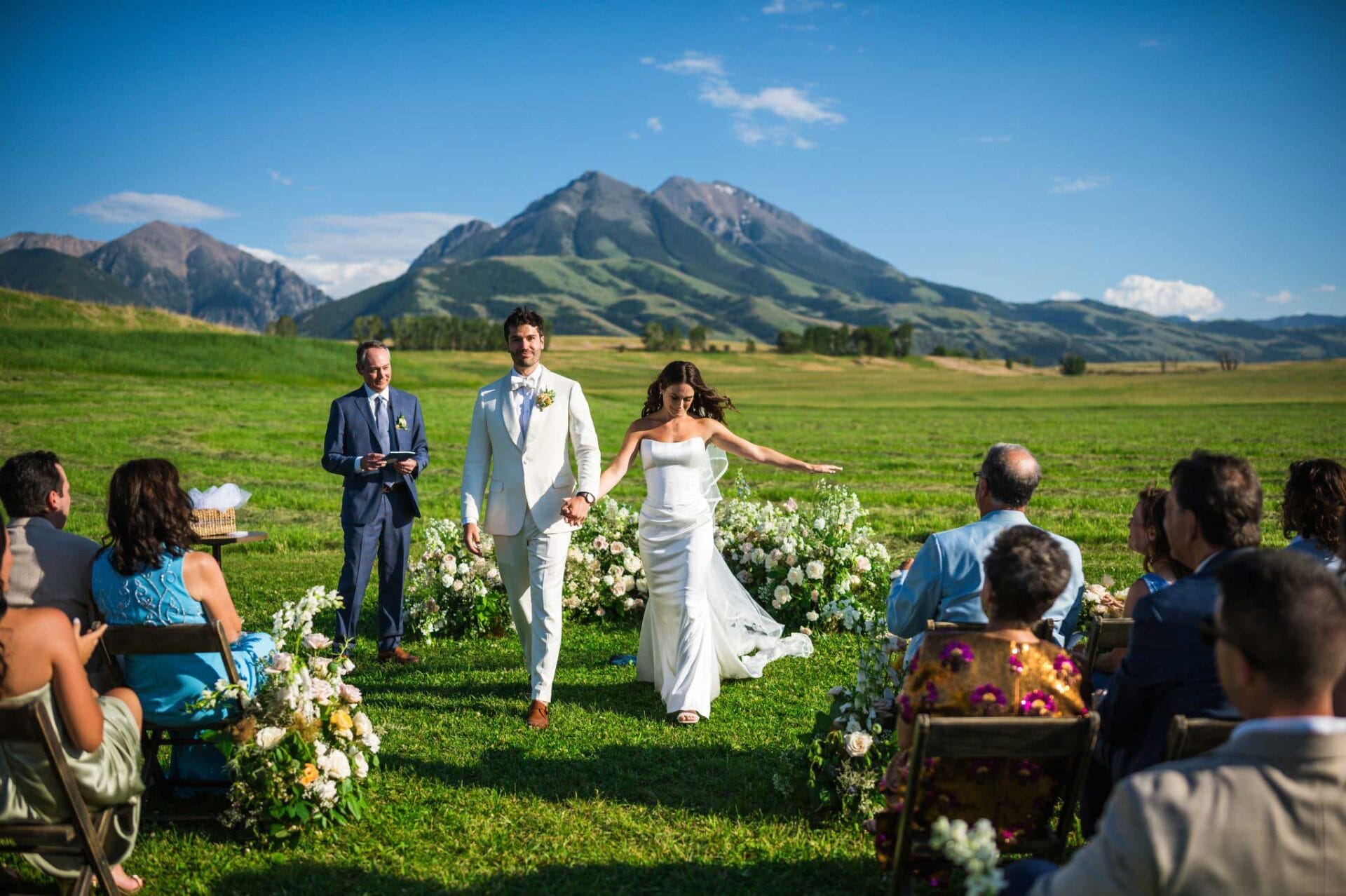 A bride and groom walk hand in hand down an outdoor aisle, with guests seated on both sides and mountains visible under a clear blue sky in the background.