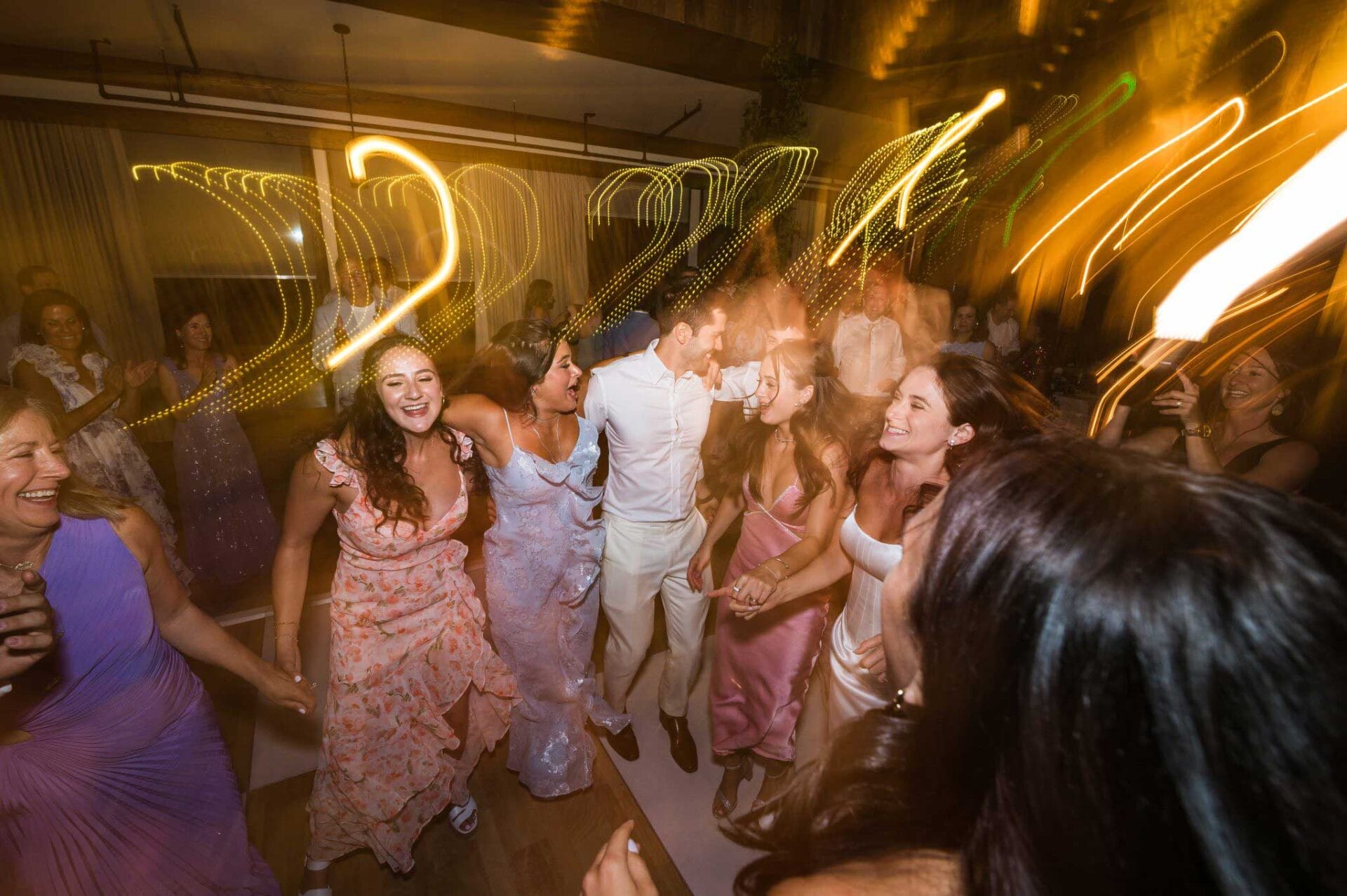 A group of people in formal attire dance energetically at an indoor event with streaks of yellow lights creating a dynamic effect.
