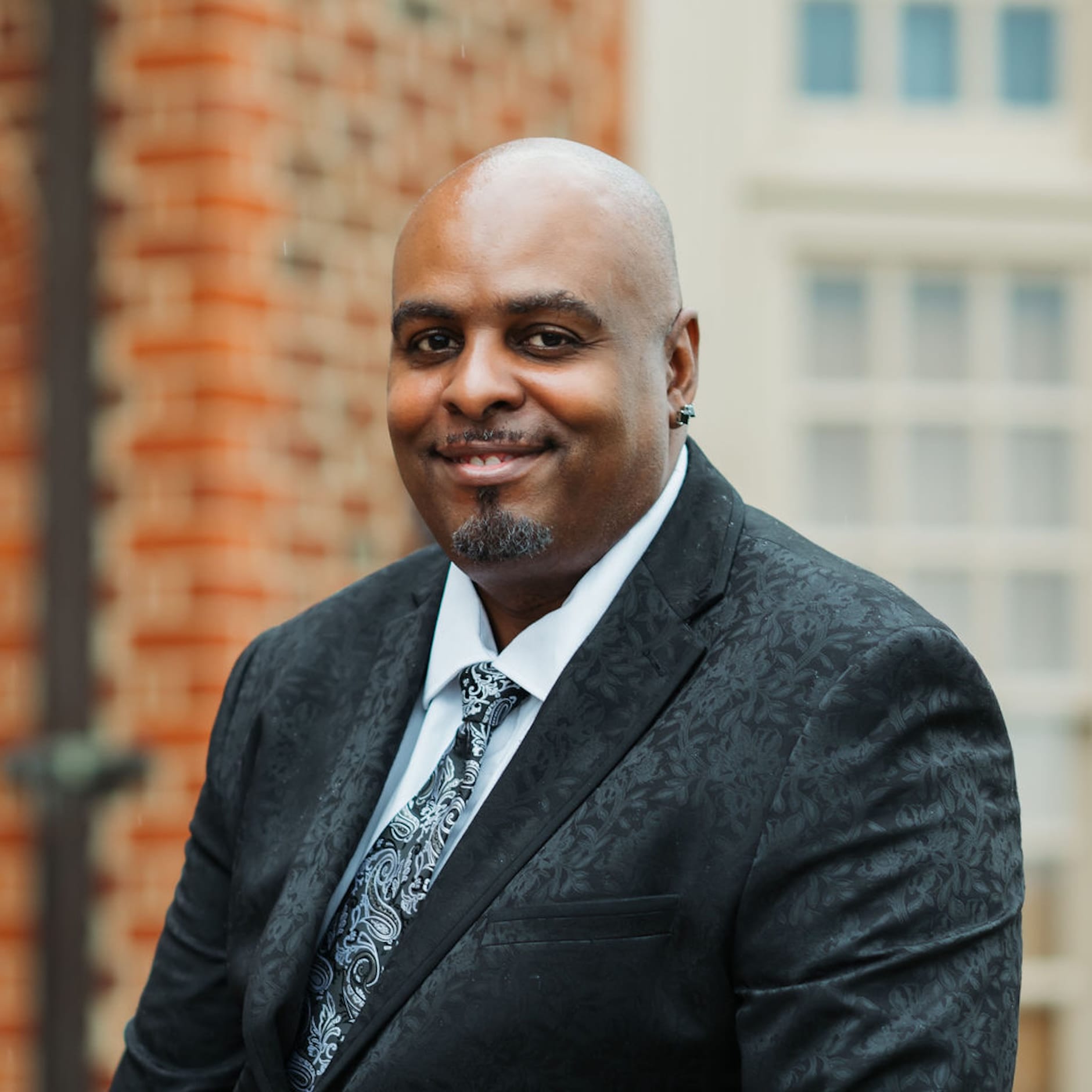 Sean Headshot A man in a patterned black suit and paisley tie smiles at the camera, standing in front of a brick wall and window.