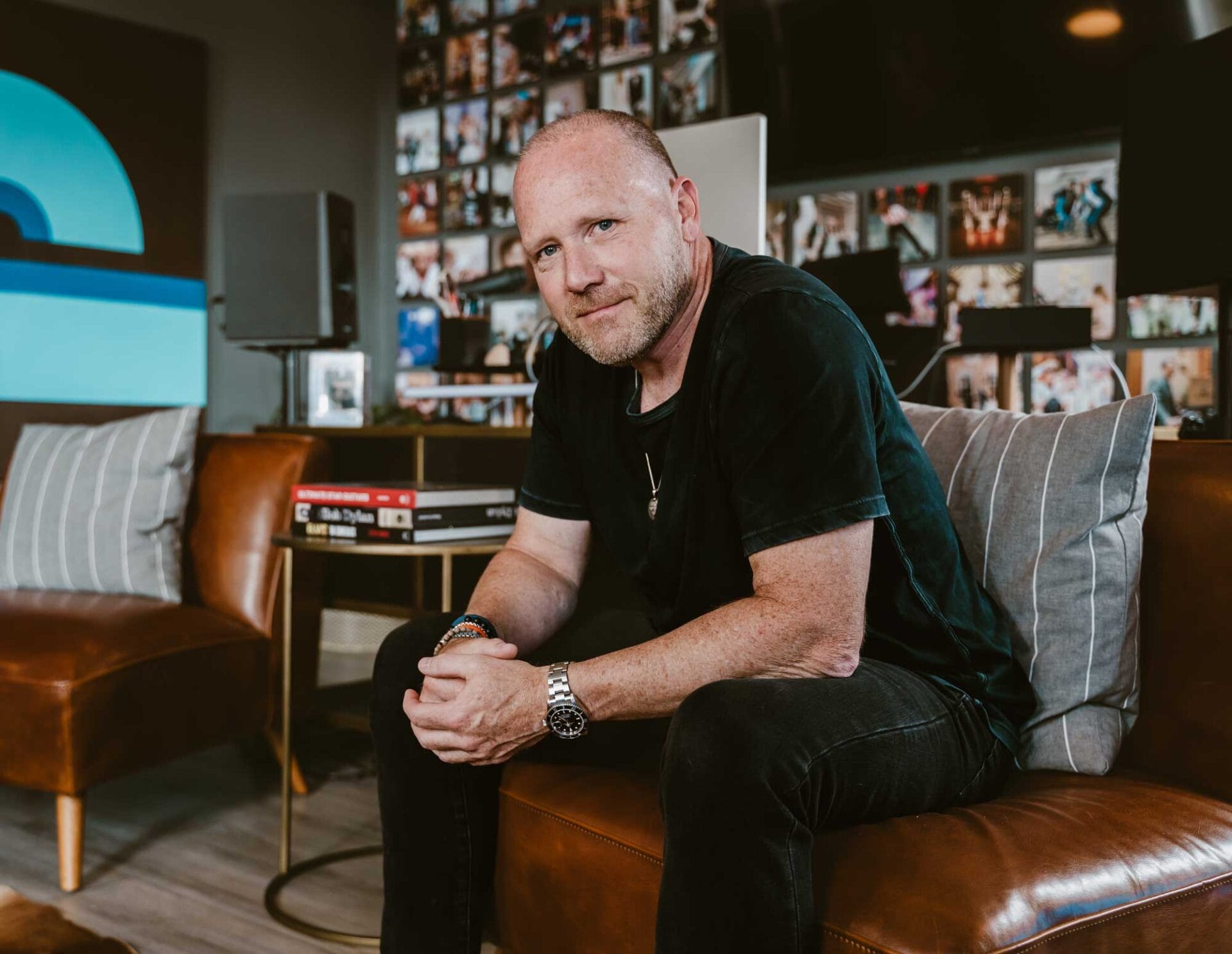 A man with a shaved head and beard sits on a brown leather couch in a modern room with photo-covered walls and contemporary decor.