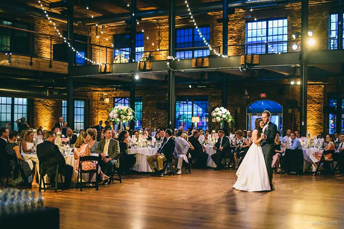 A bride and groom share their first dance in the center of a decorated, industrial-style reception hall as guests seated at round tables watch.