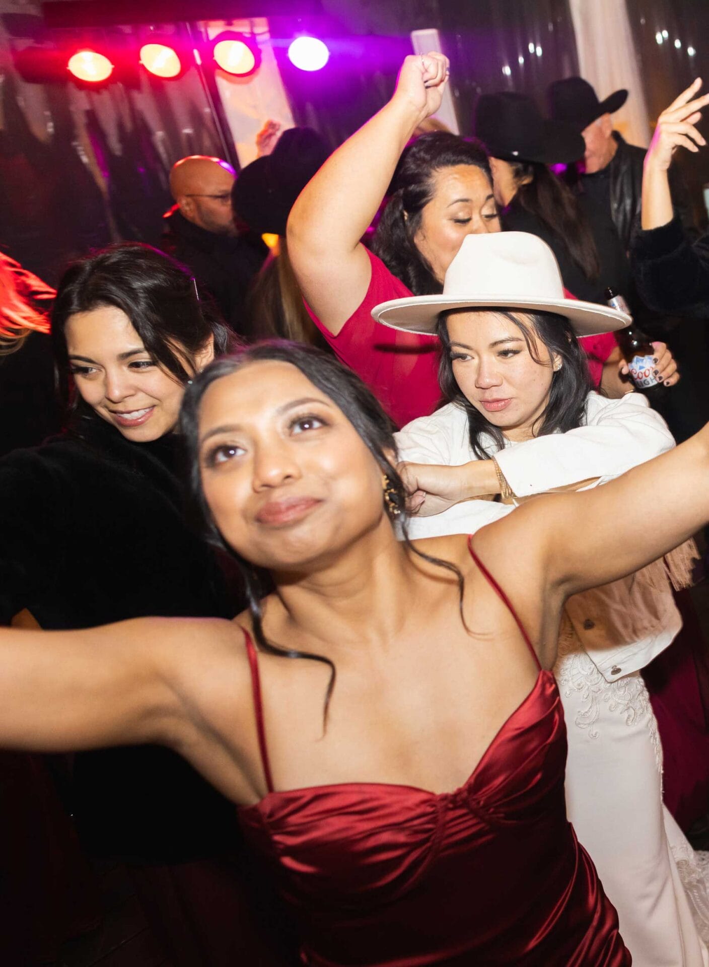 A group of people dance and celebrate at a party, with colorful lights in the background. The focus is on a woman in a red dress smiling with arms raised.