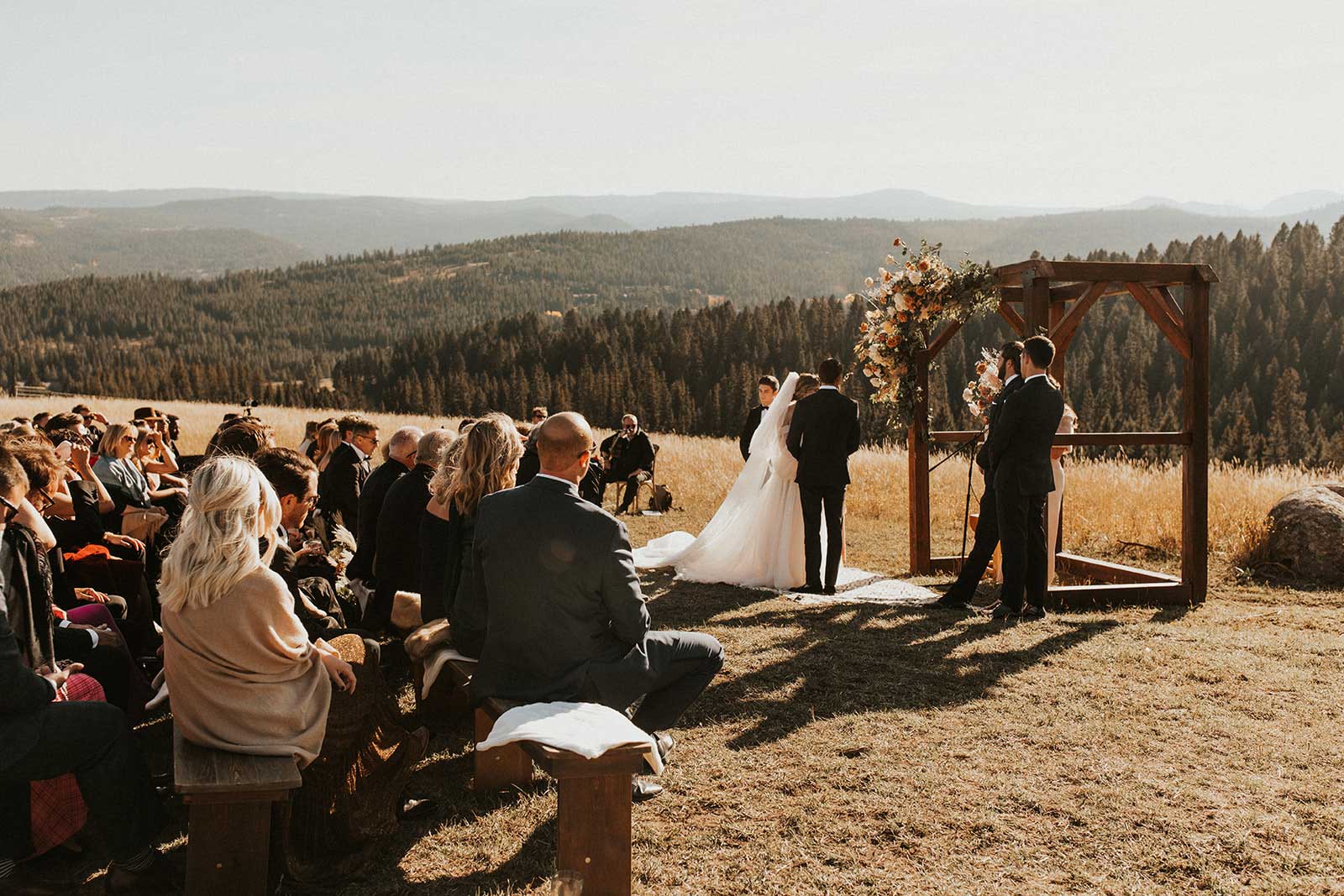 Outdoor wedding ceremony in a field with a wooden arch, bride and groom facing the officiant, and guests seated on benches; mountains and forest in the background.