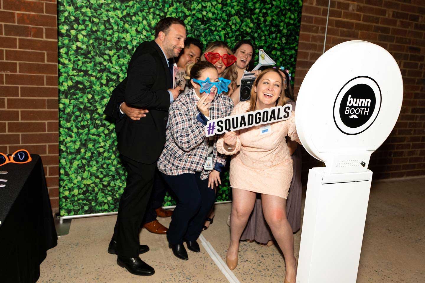 A group of people pose together in front of a photo booth, holding props and a sign that reads "#SQUADGOALS," with a green leafy backdrop and a bunn booth photo station.