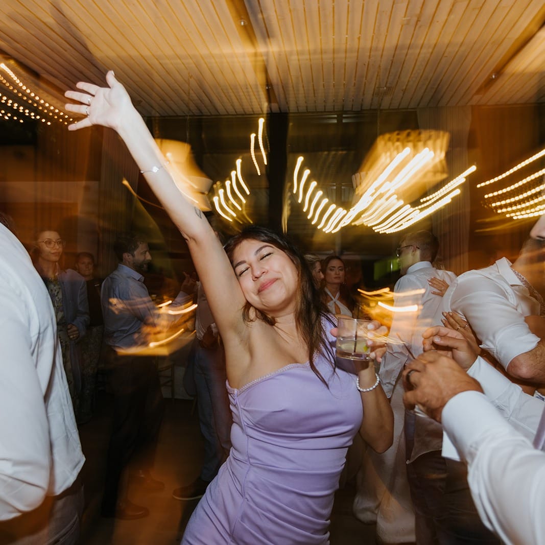 bunn charleston dj girl dancing Woman in a lavender dress dances with one arm raised, holding a drink, surrounded by people with blurred lights in the background.