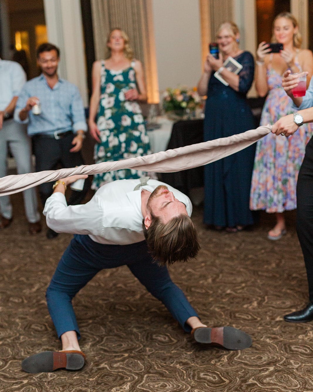 bunn charleston dj guy doing limbo A man in formal attire plays limbo under a cloth while people in semi-formal dresses and suits watch and take photos at an indoor event.