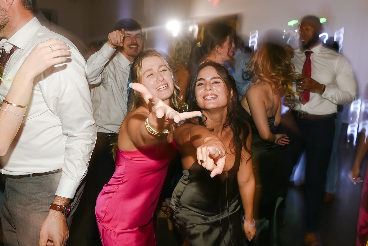 bunn charlotte dj girls dancing with their hands out front Two women in formal dresses smile and reach toward the camera while dancing at a party, surrounded by other people in festive attire.