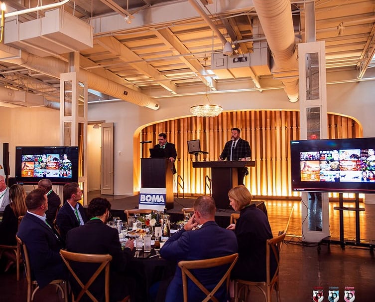 Two speakers stand at podiums in a modern event space, addressing seated attendees; two large screens display images beside them.