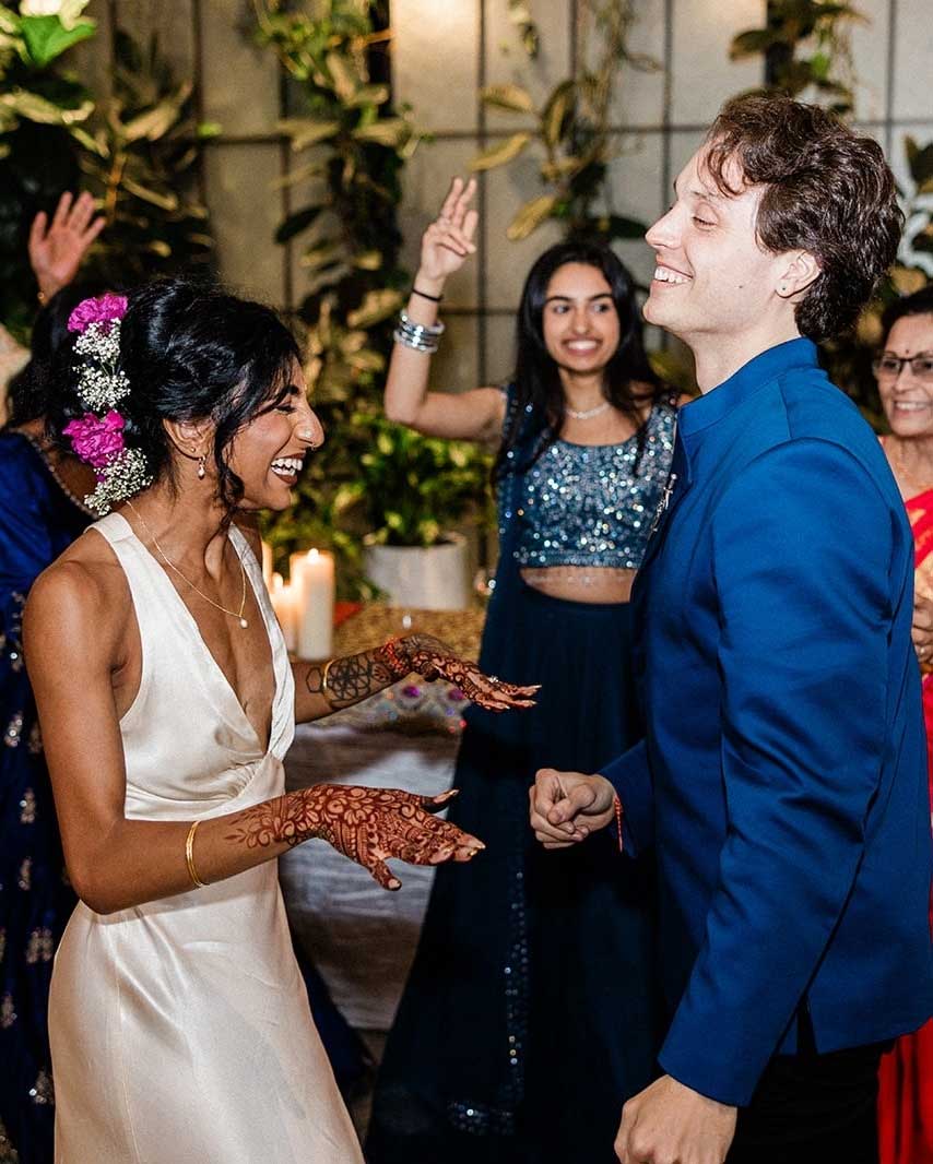 bunn-dj-indian-wedding-san-diego A woman in a white dress with henna on her hands and a man in a blue suit smile and dance at a festive indoor event with others in the background.