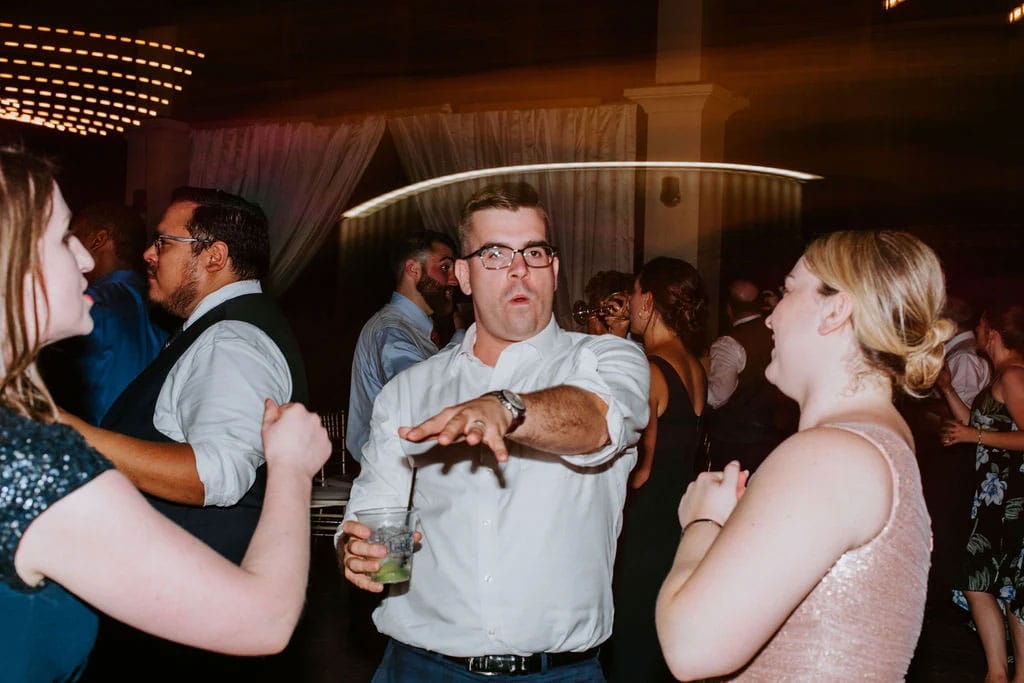 A man holding a drink gestures animatedly while dancing with others at an indoor event, surrounded by people in formal attire.