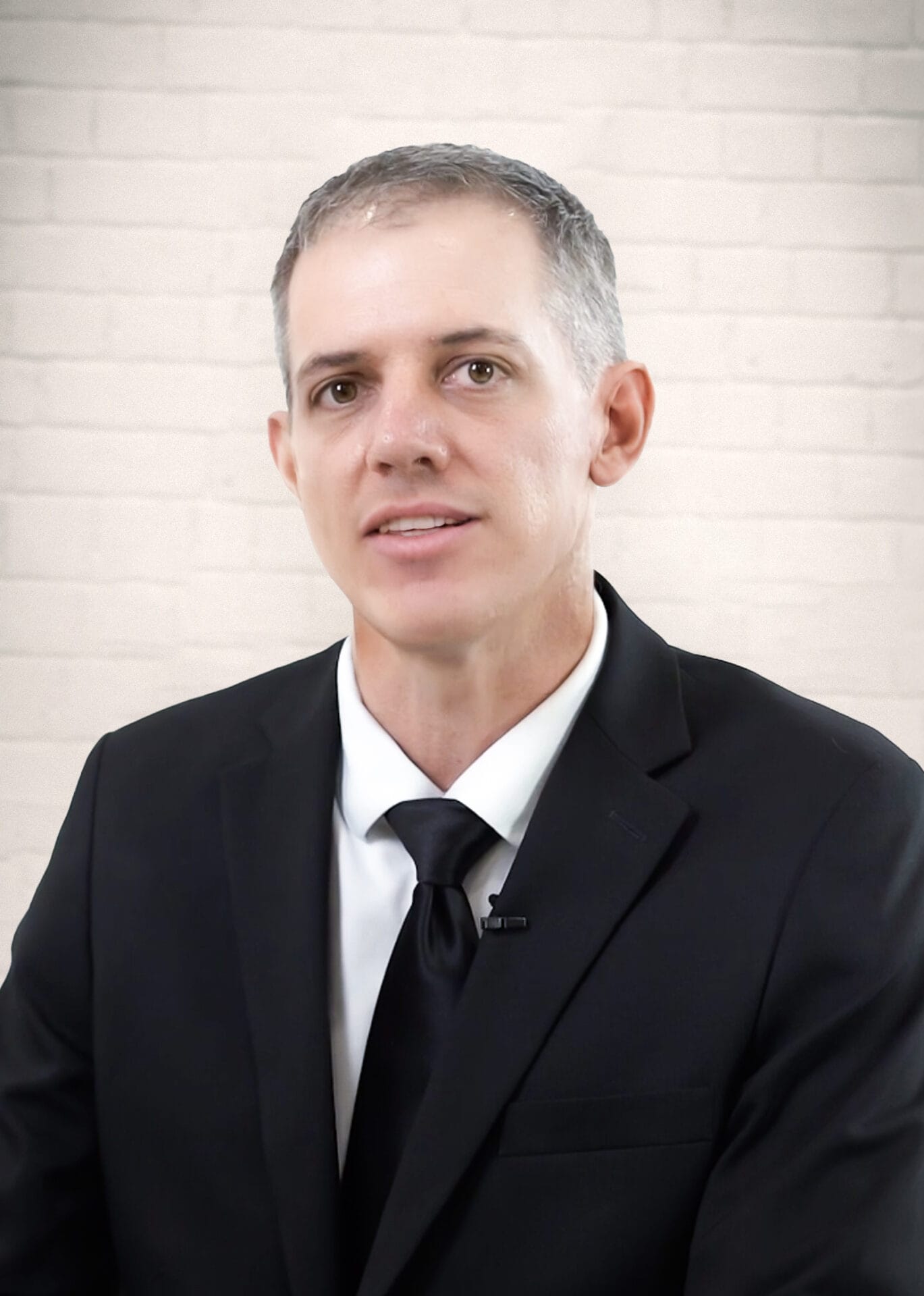 A man in a black suit and tie sits in front of a light-colored brick wall, looking at the camera.