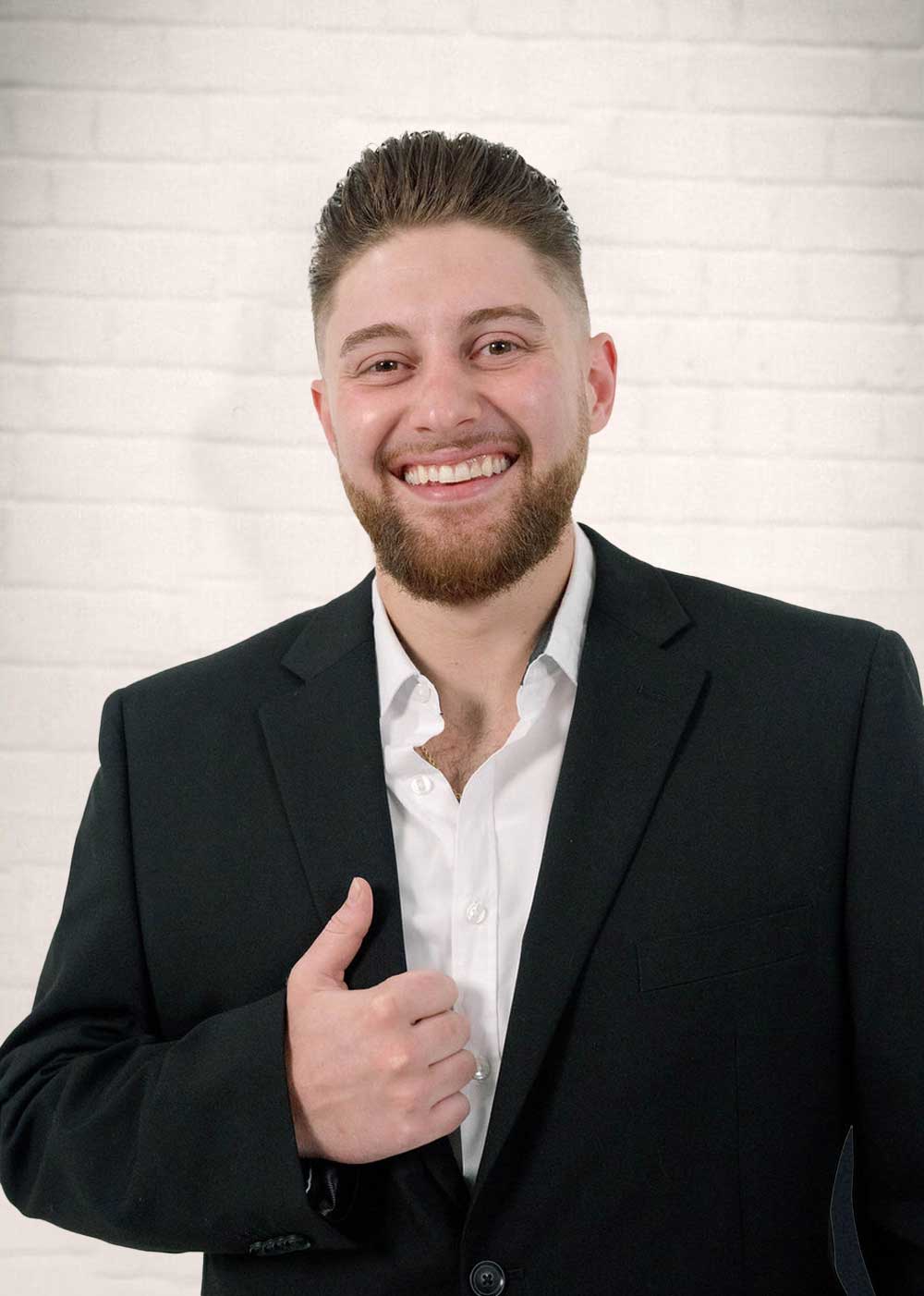 A man with short hair and a beard smiles at the camera, wearing a black suit and white shirt, giving a thumbs-up gesture in front of a white brick wall.