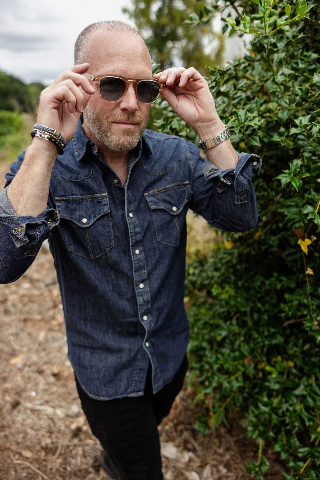joe-bunn-denim-nature-sunglasses A man in a denim shirt adjusts his sunglasses while standing outdoors next to green foliage.