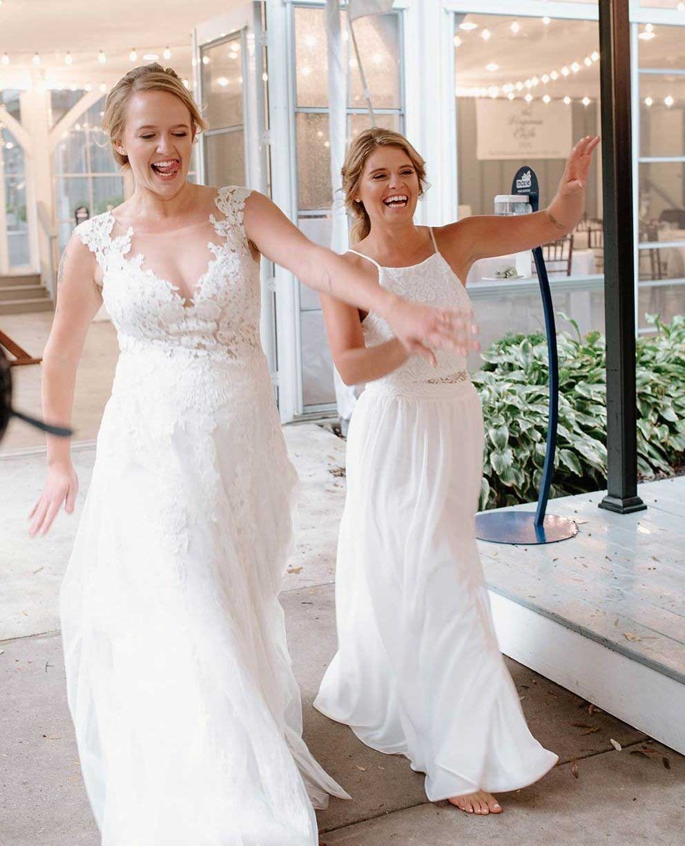 lgbtq-richmond-wedding Two women in white dresses, one barefoot, smile and extend their arms while walking outside near a decorated venue with string lights.