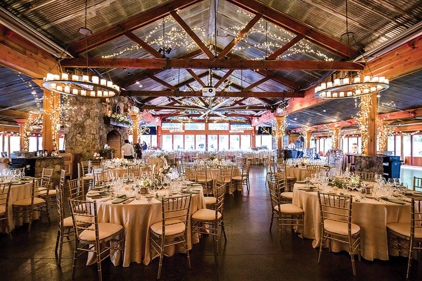 Large banquet hall with round tables set for an event, featuring gold chairs, white tablecloths, and string lights on wooden beams—perfect for those seeking elegant Raleigh Wedding Venues.