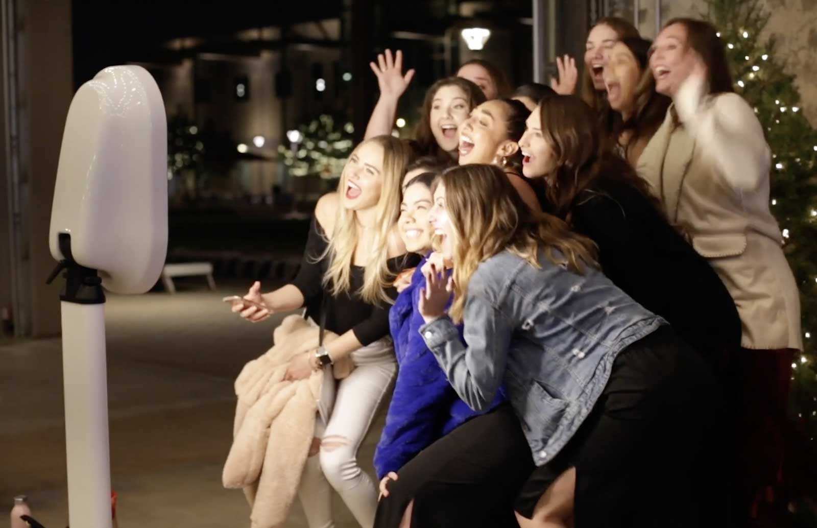 A group of women poses and smiles enthusiastically for a photo booth at an indoor event, with lights and greenery in the background.