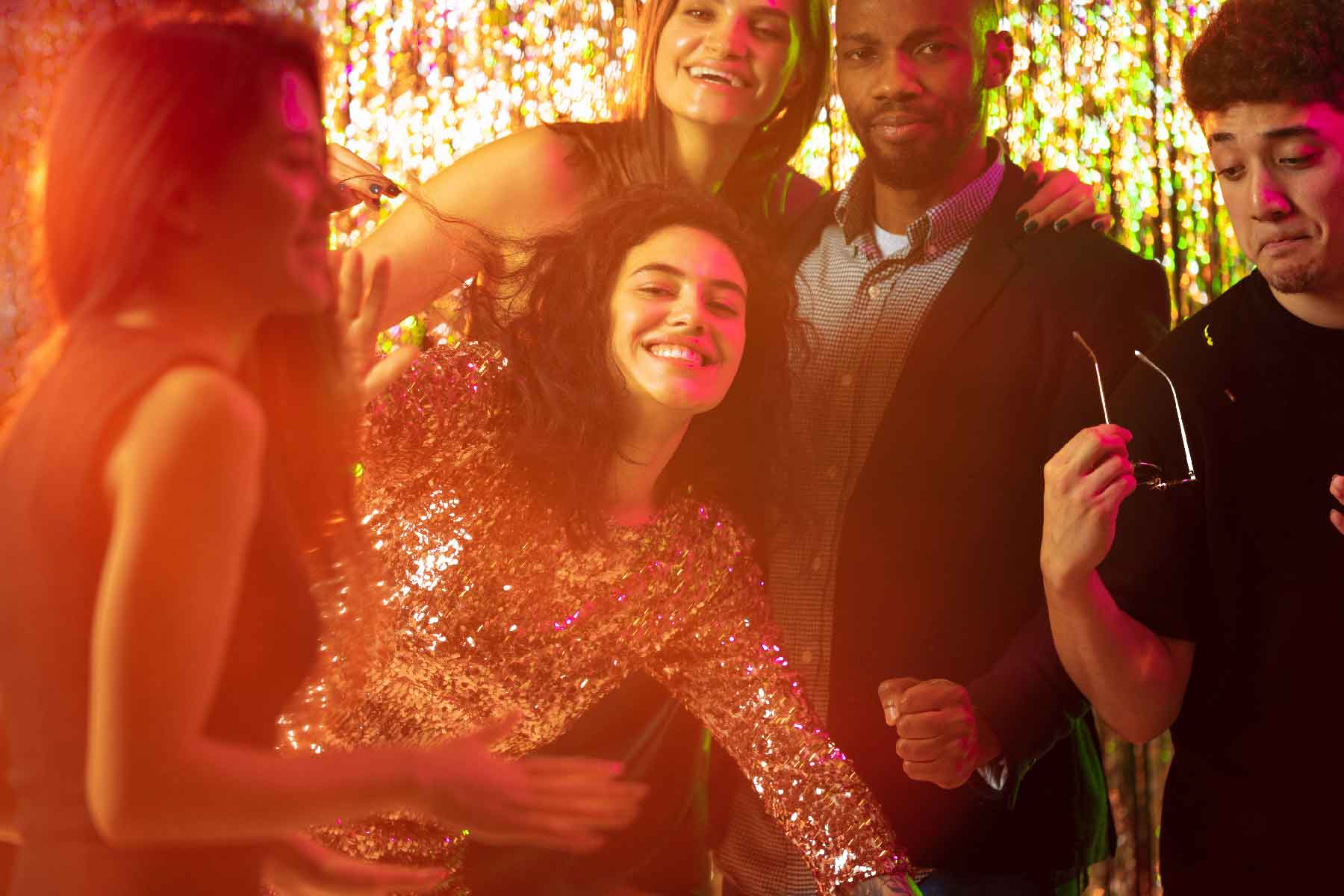 Five young adults stand close together at a party, smiling and laughing, with sparkling gold decorations in the background.