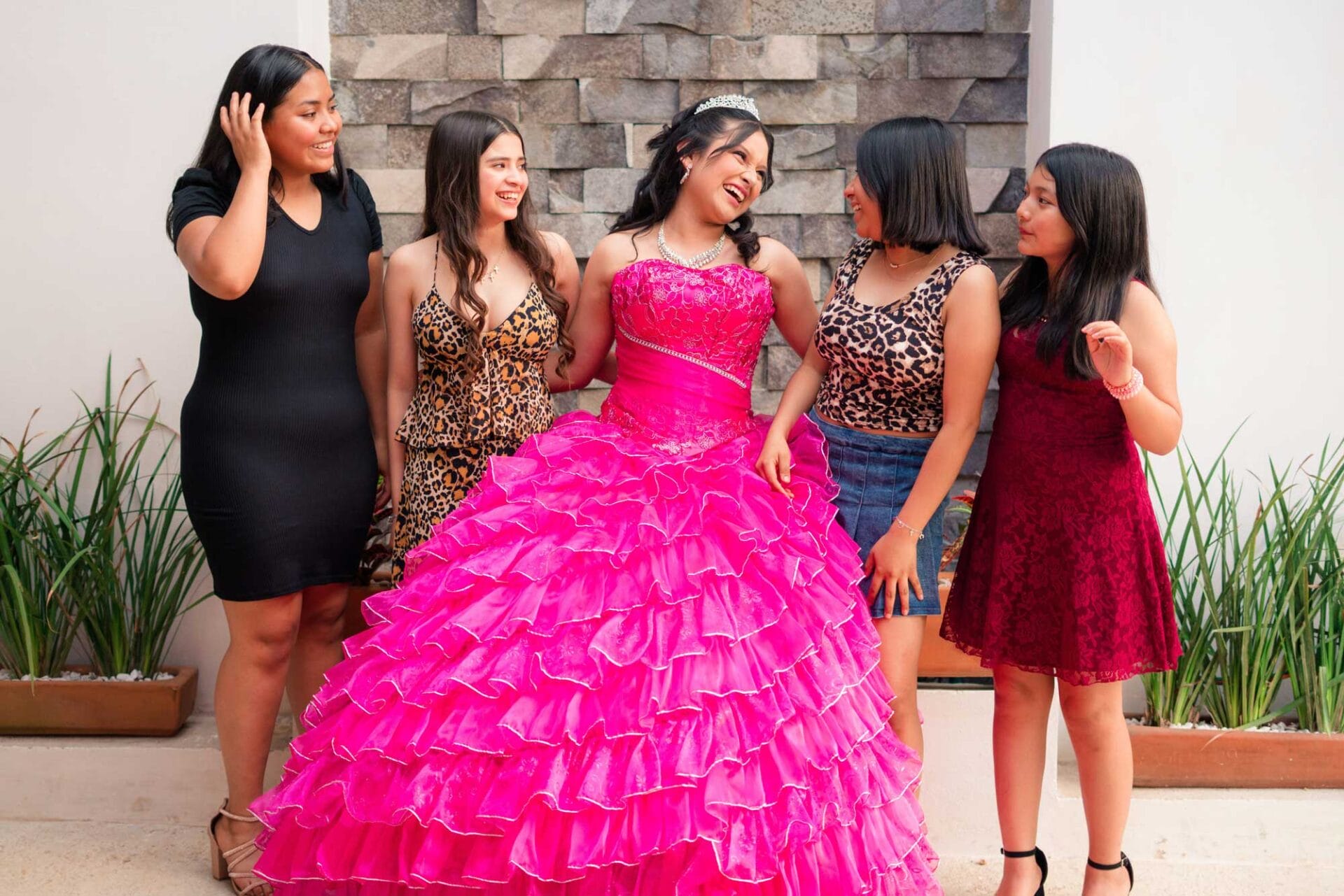 quinceanera-girl-with-friends Five young women pose together outdoors; one wears a bright pink, ruffled ball gown and a tiara, while the others wear casual or semi-formal dresses, standing in front of a stone wall.