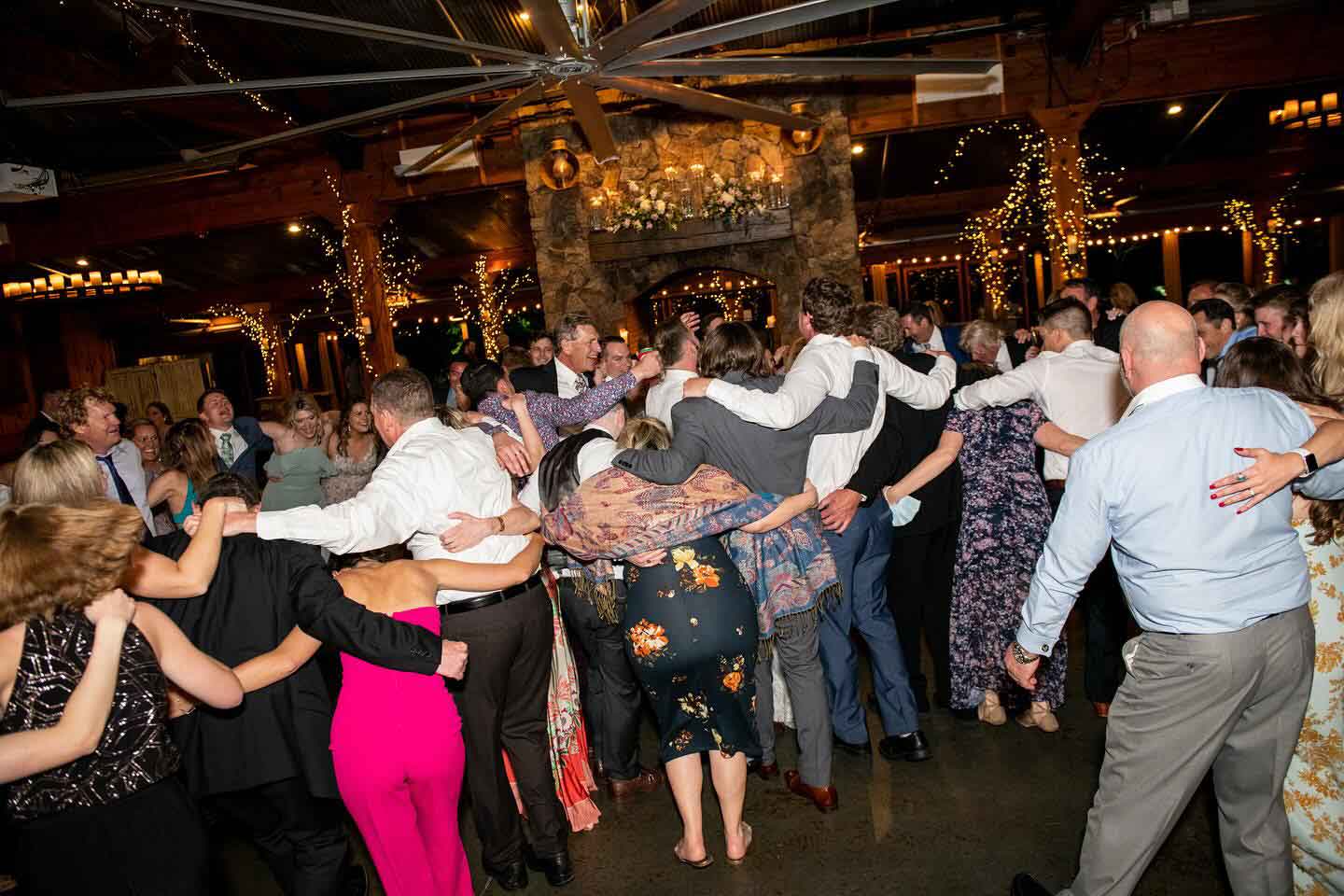 A large group of people stand in a circle on a dance floor with arms around each other at an indoor event, with string lights and a stone fireplace in the background.