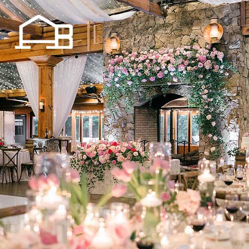 A rustic event space with stone walls, wooden beams, and a fireplace decorated with pink flowers and greenery; tables are set for a formal gathering.