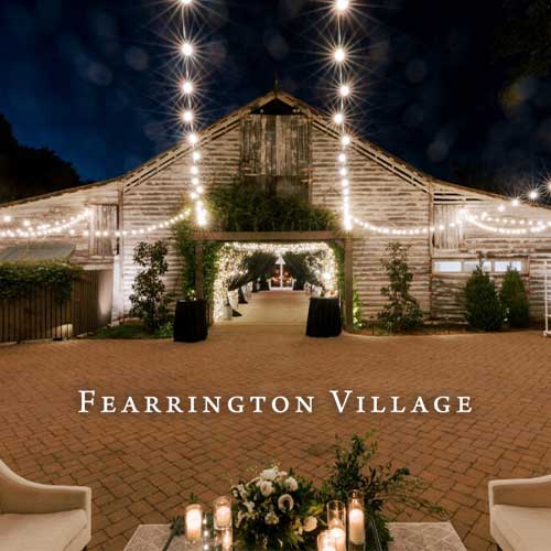A rustic barn at Fearrington Village is lit with string lights at night, with a brick patio and floral centerpiece in the foreground.