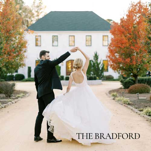 A bride and groom dance outside on a dirt path in front of a white building with autumn trees; text reads “The Bradford.”.