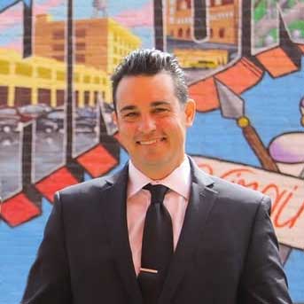 richmond-bunn-dj-owner-frank A man in a dark suit and tie stands smiling in front of a colorful mural featuring city buildings and large text.